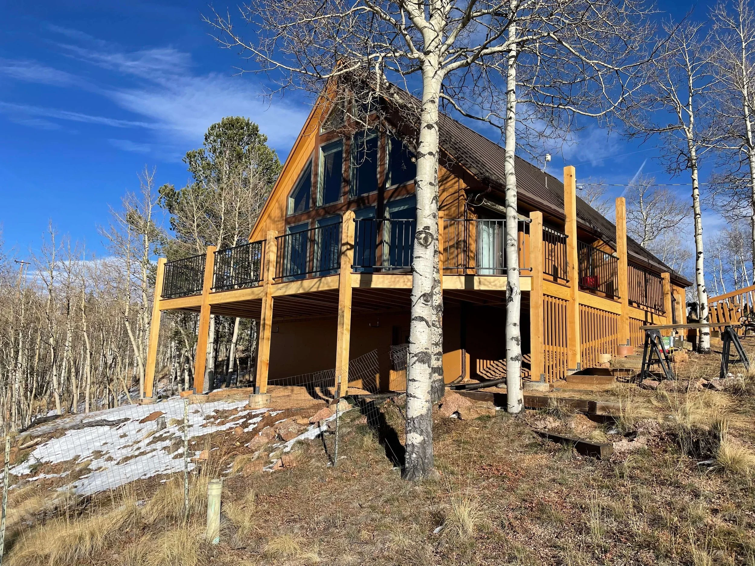 A wooden house with large windows and a steep roof, surrounded by leafless trees and a partly cloudy blue sky, with some snow on the ground.