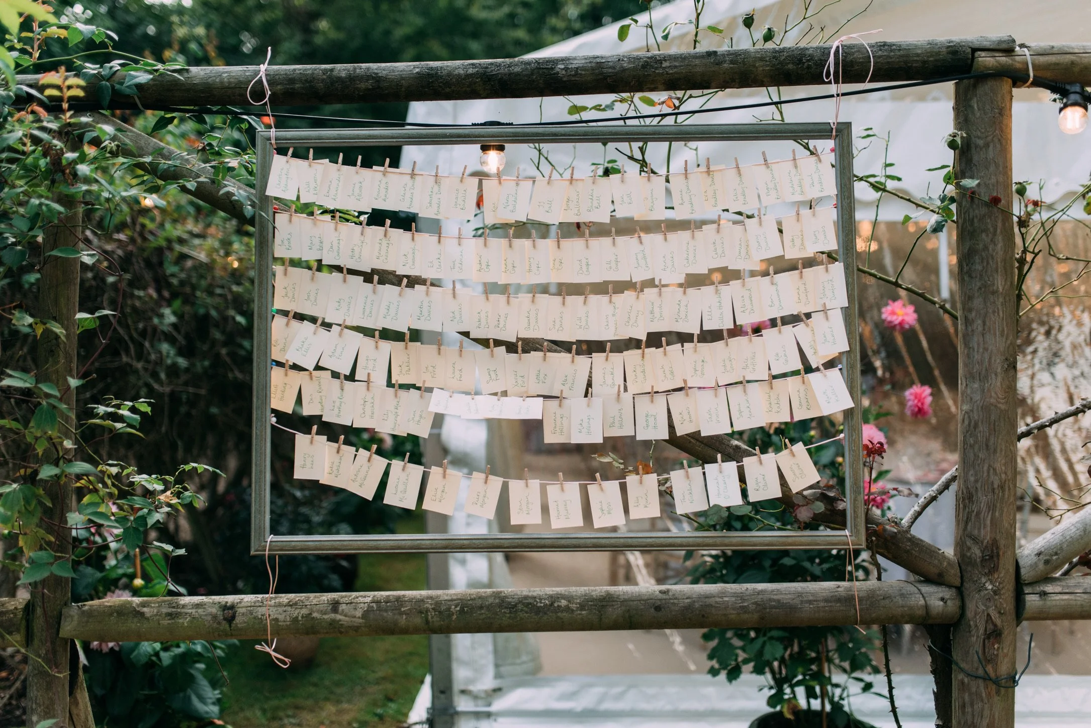 Multiple rows of white paper cards hung with clothespins on strings, displayed on a wooden frame outdoors, possibly for a wedding or event seating arrangement.