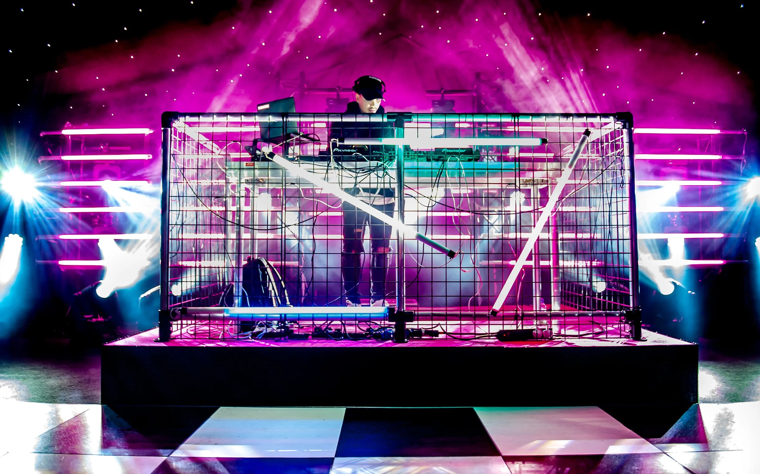 A DJ performs on a stage with vibrant pink, purple, and blue neon lights, surrounded by a metal cage and a checkerboard floor.