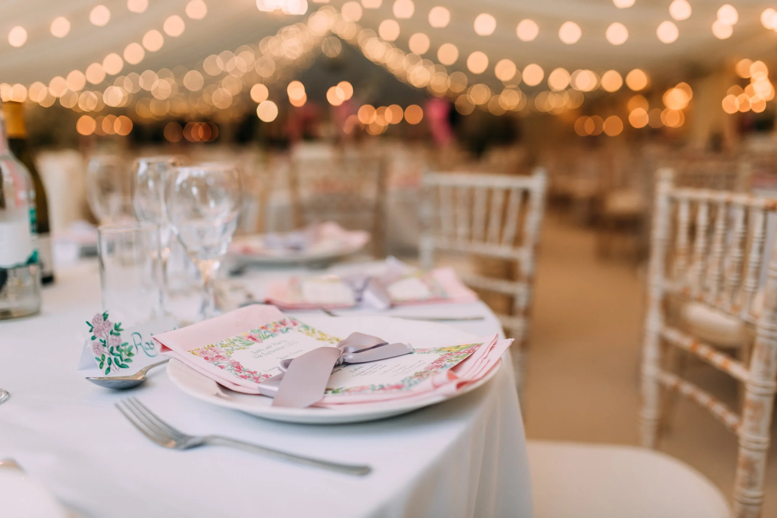 Elegant wedding reception table decorated with pink napkins, silver utensils, wine glasses, and floral place cards, with a blurred background of string lights and chairs.