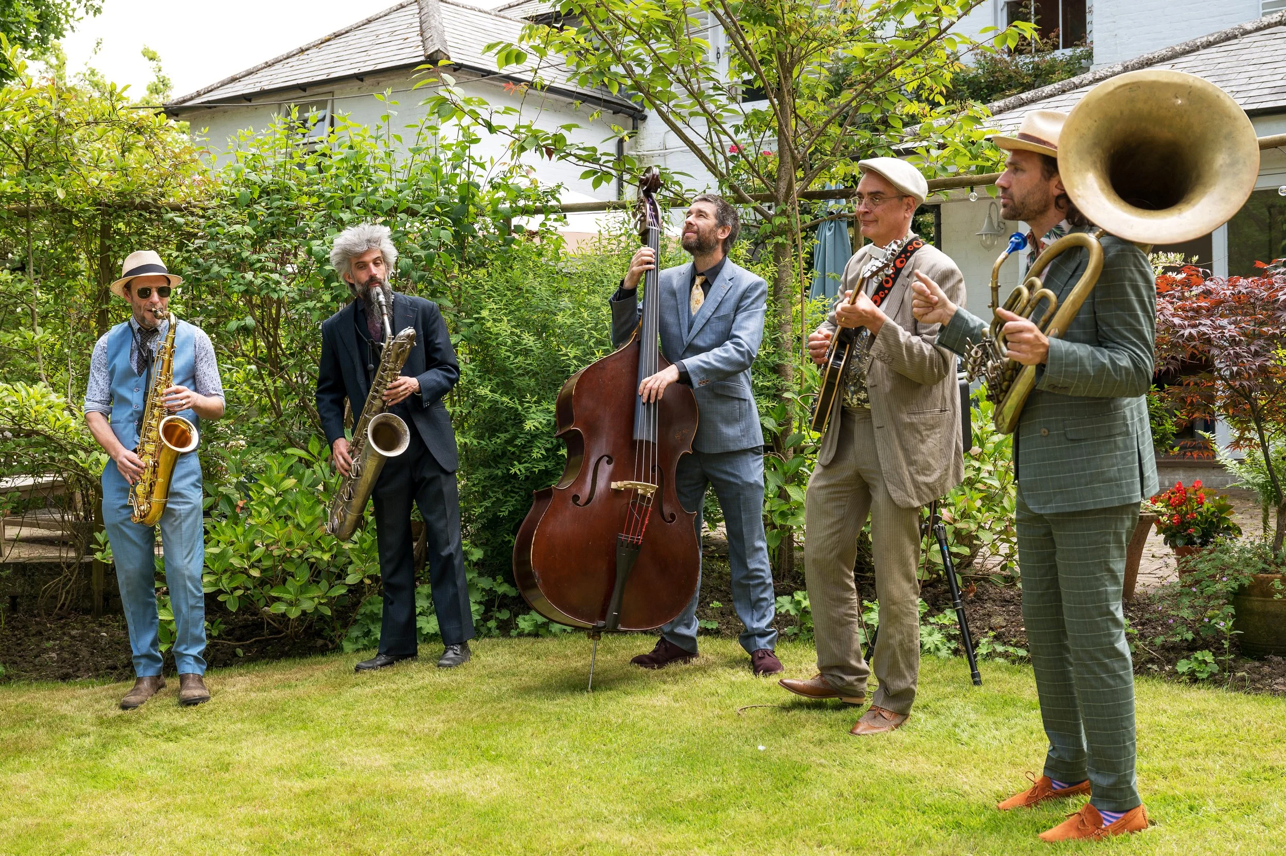 A band of five musicians playing instruments outdoors in a garden with green foliage, a tree, and a house in the background. The musicians are playing saxophone, clarinet, double bass, electric guitar, and tuba, dressed in vintage-style clothing.