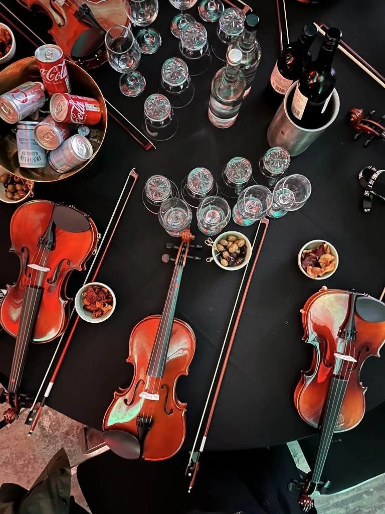 Table with three violins, several empty wine glasses, cans, bottles of wine, bowls of snacks, and chopsticks.