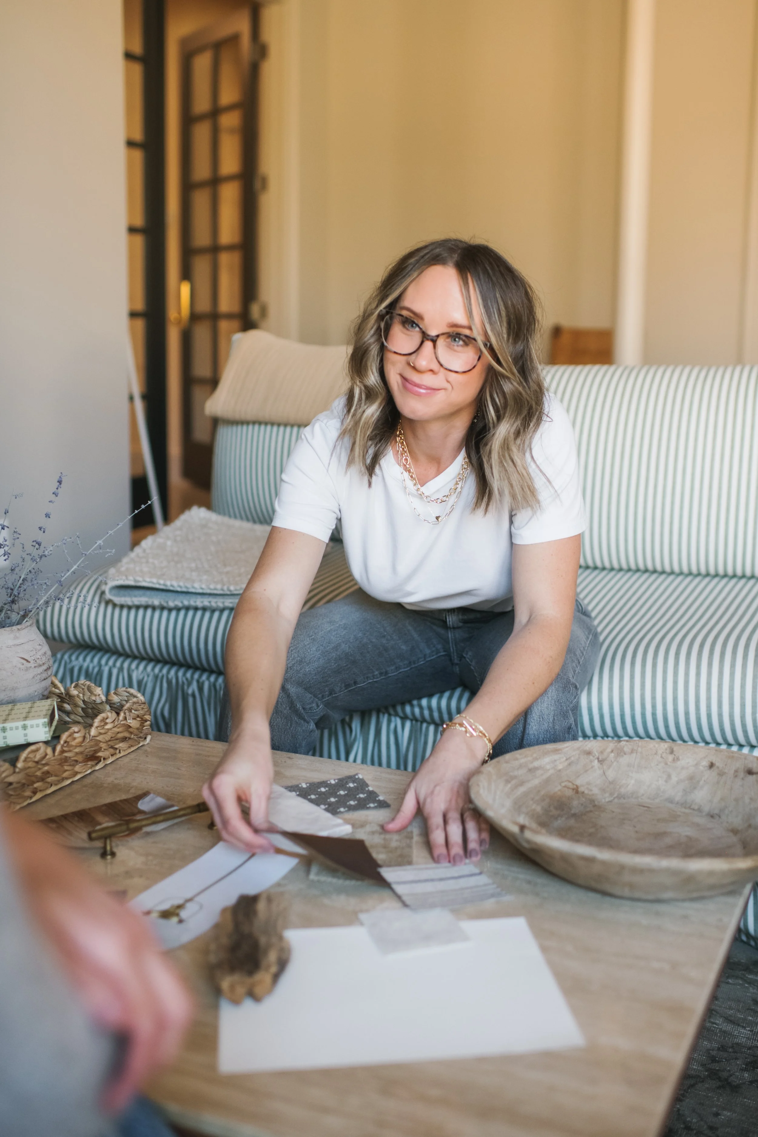 A portrait of interior designer, Trista Cross. She's wearing glasses and curled hair sitting on a striped sofa, working on a creative project at a wooden table.