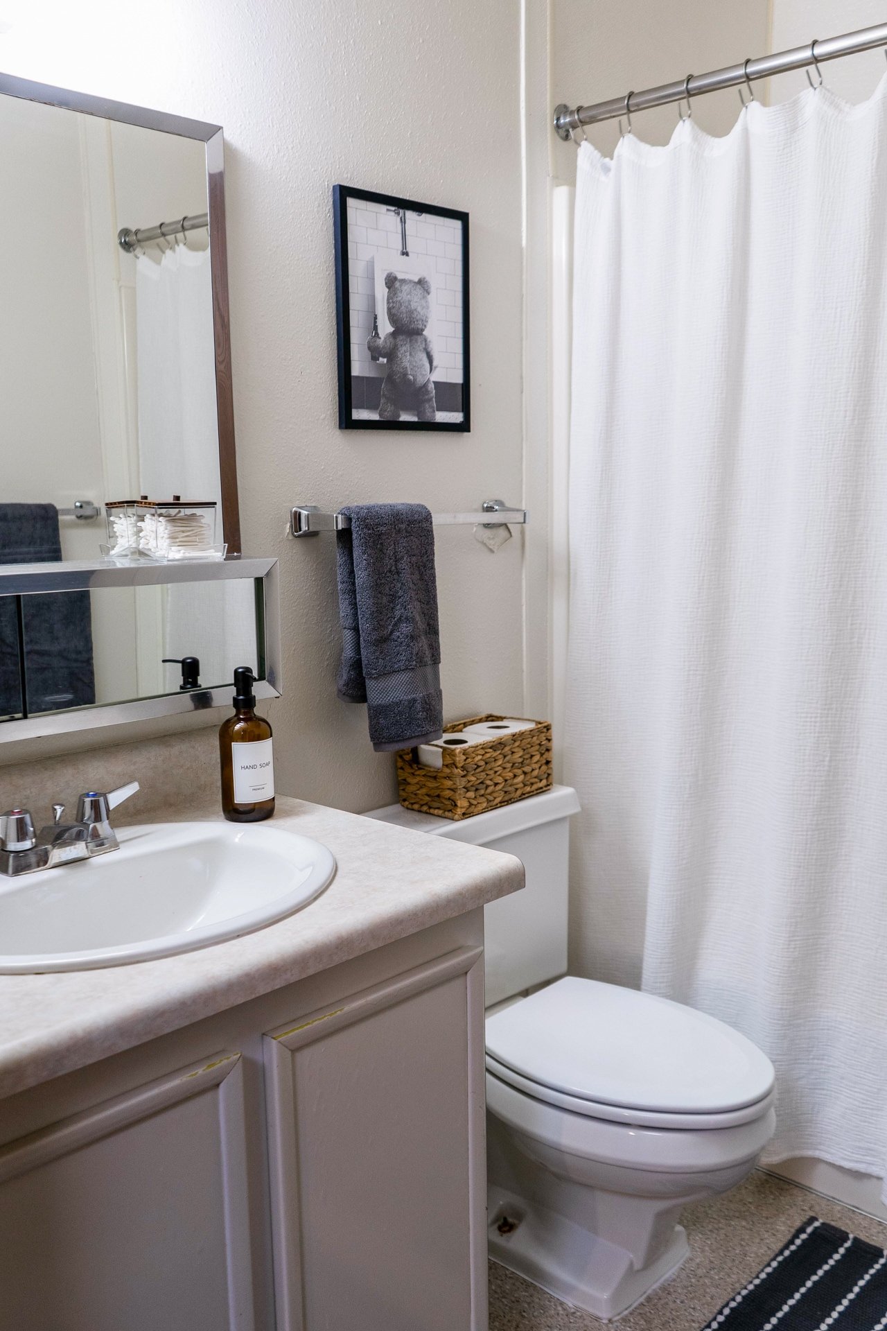 Small bathroom with a white sink, brown soap dispenser, a mirror, a gray towel on a towel rack, a framed black-and-white picture of a bear in a shower, a basket on top of the toilet, and a white shower curtain.