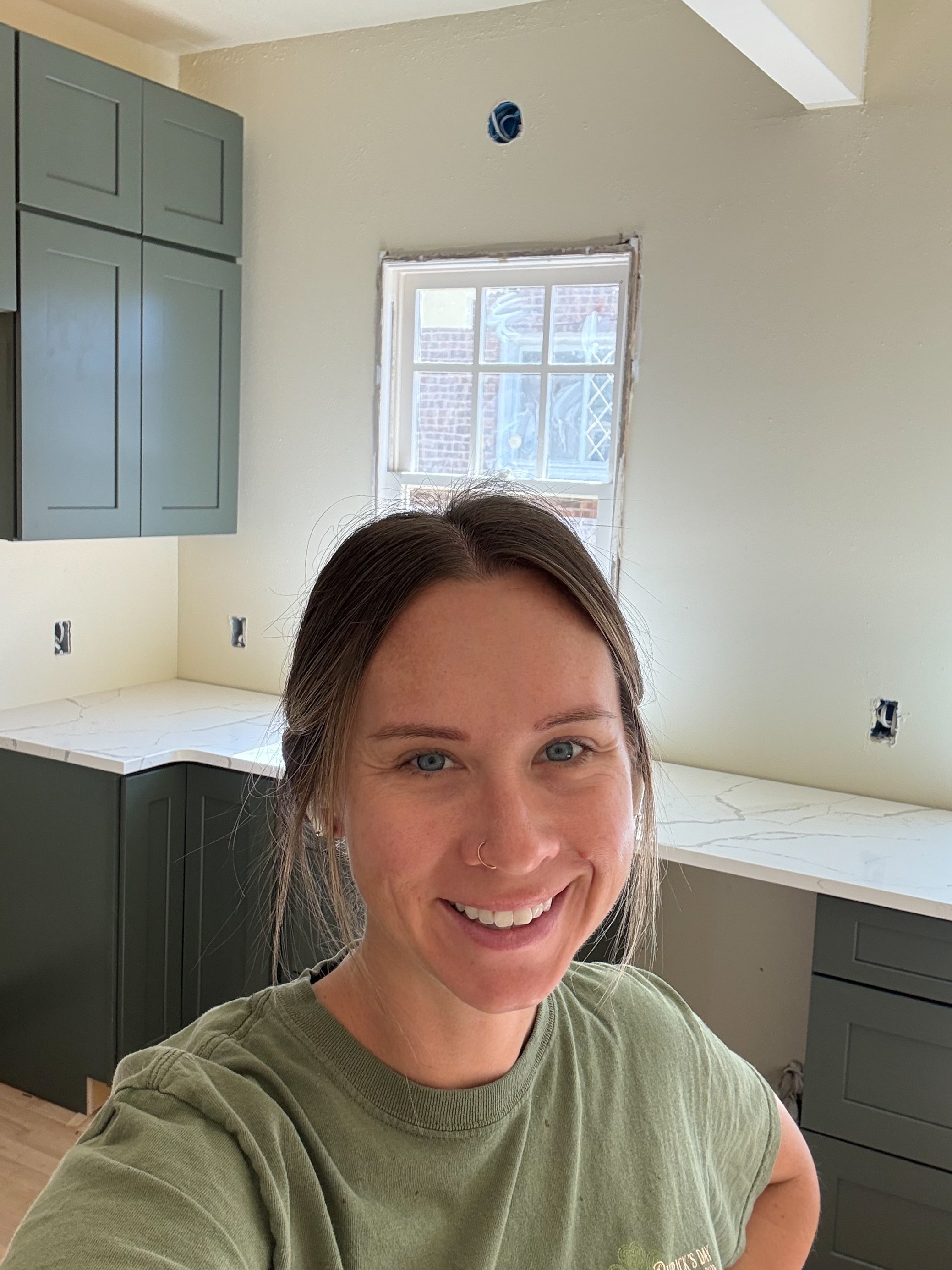 Interior designer, Trista Cross, with brown hair, blue eyes, and a gold nose piercing smiling in a kitchen under renovation, with green cabinets, a marble countertop, and a window in the background.