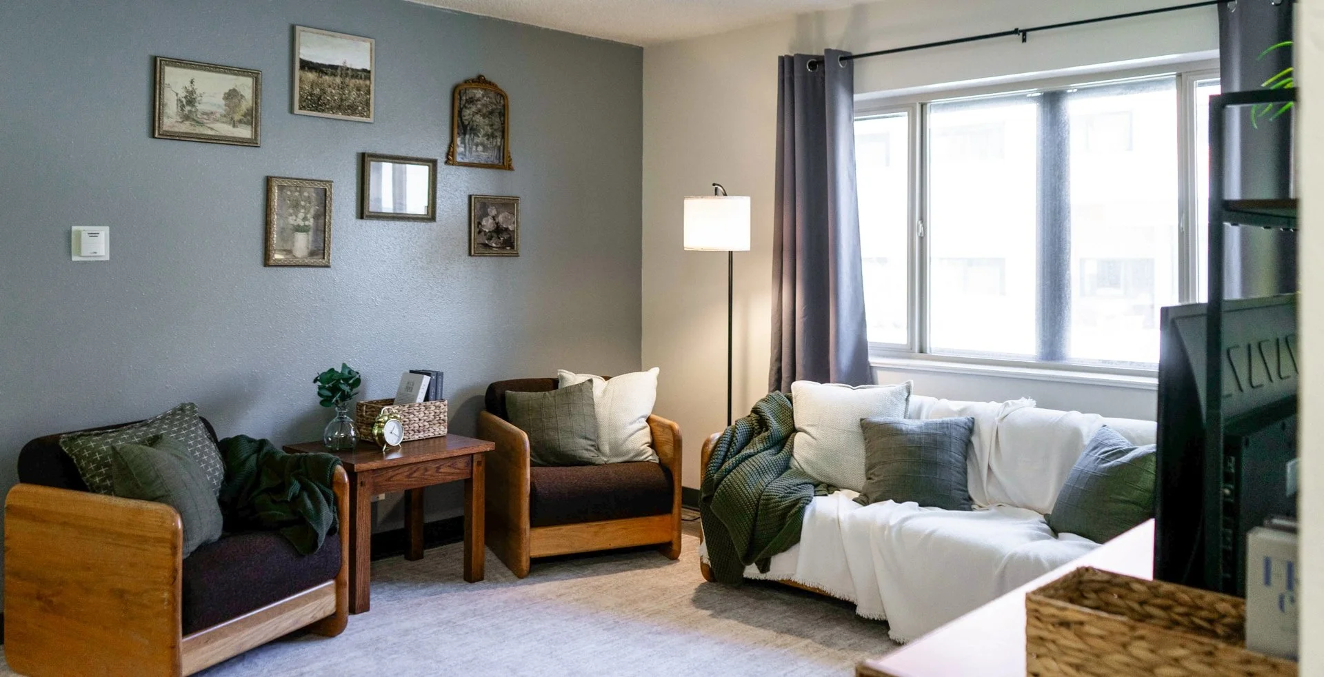 Living room with cushioned chairs, a white sofa with pillows, a floor lamp, wall art, and a large window with dark curtains.