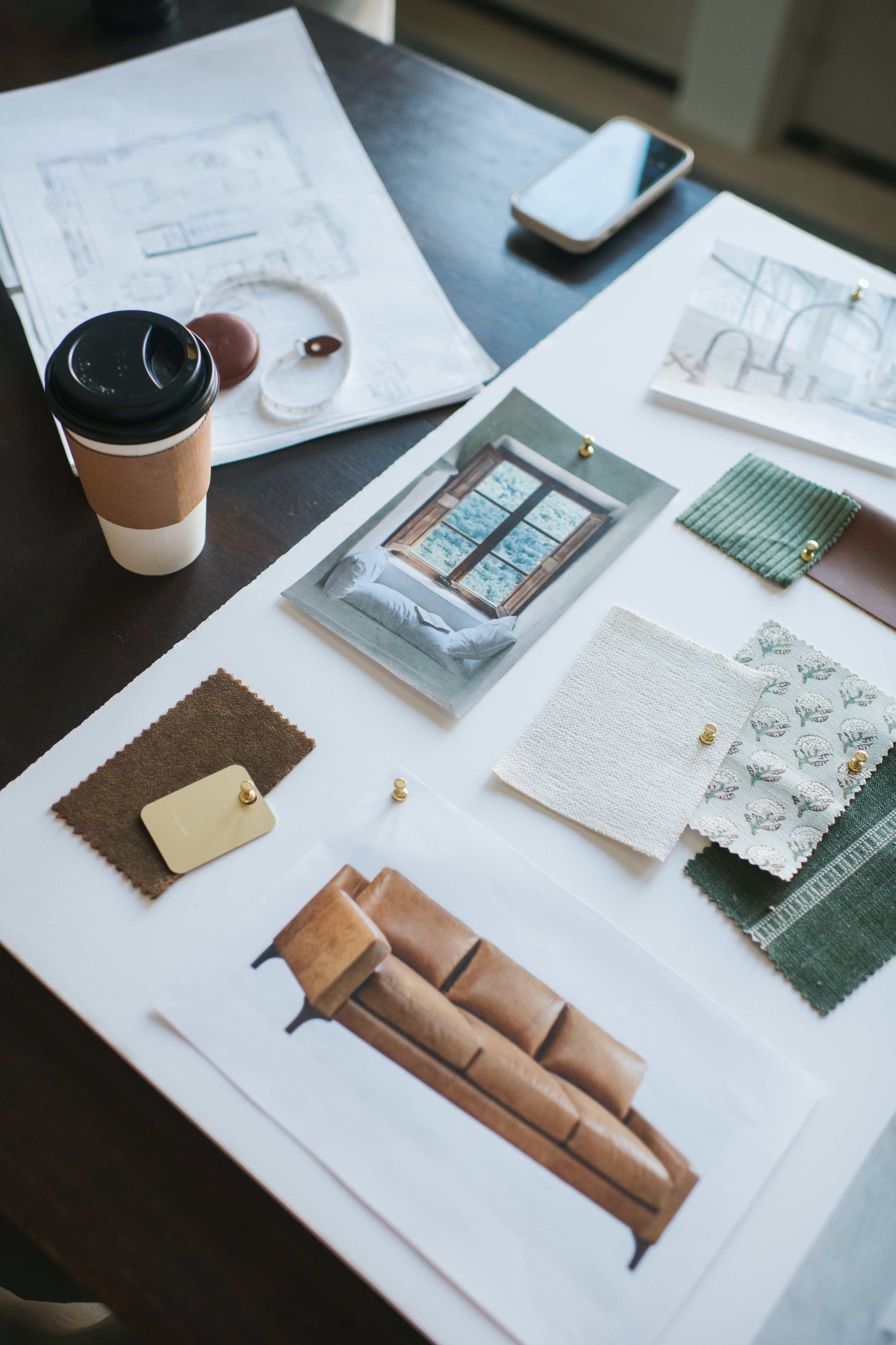 Interior design workspace with fabric samples, color swatches, design photos, moodboard inspiration, a coffee cup, and a smartphone on a dark table.