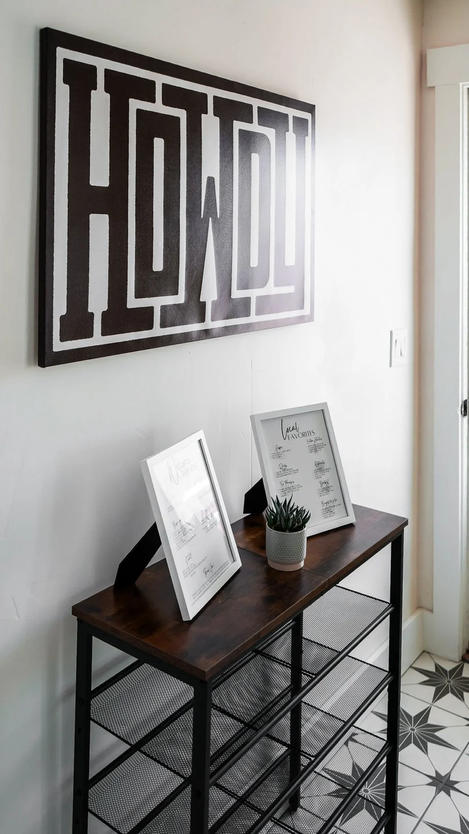 A black and white wall art piece with the word 'HOWDY' in bold, stylized letters hangs above a wooden console table. The table has two framed menus or lists, and a small potted plant. The floor is decorated with patterned black and white tiles.