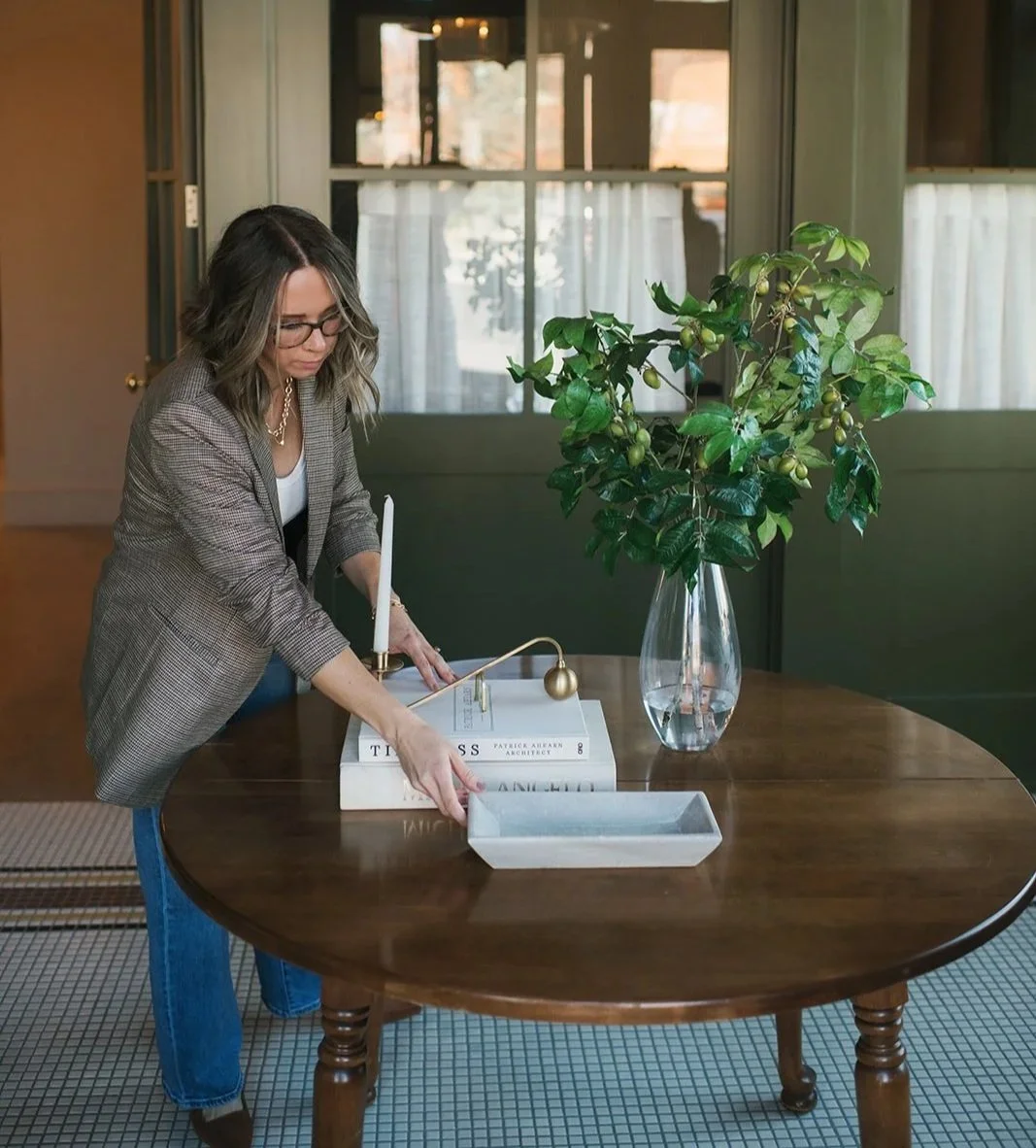 Woman arranging decorative items on a round wooden table inside a room, with books, a tall vase with a green leafy plant, and a modern table lamp.