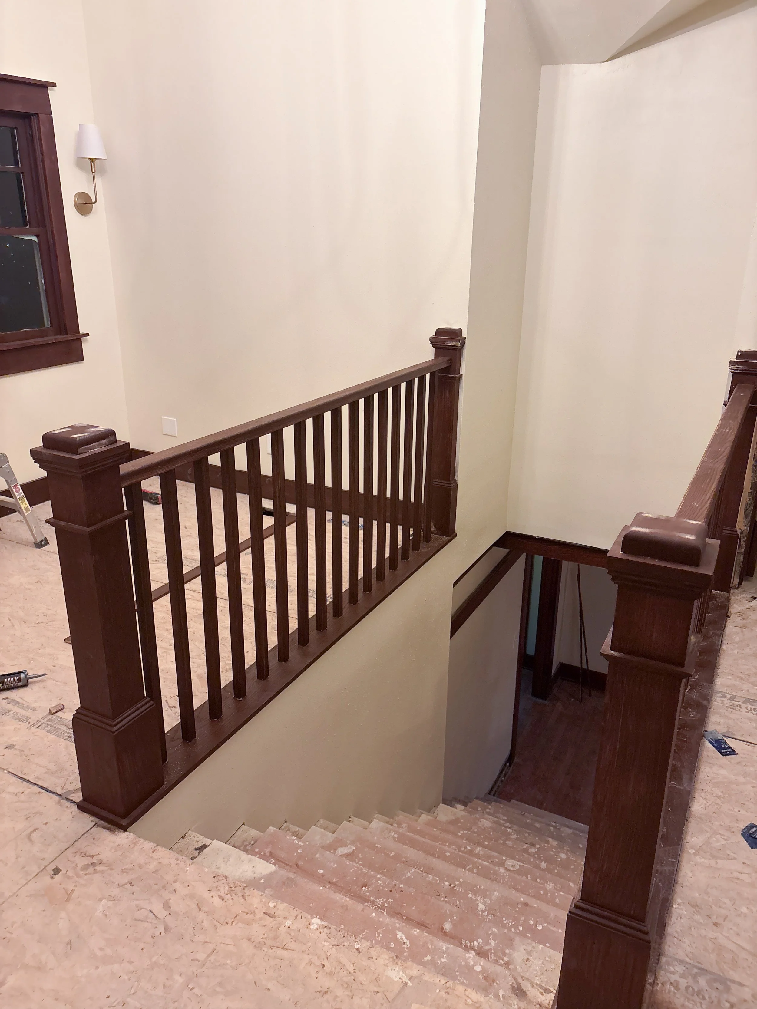Interior view of a staircase with wooden railing and newel posts, unfinished flooring, and walls, in a house under renovation.