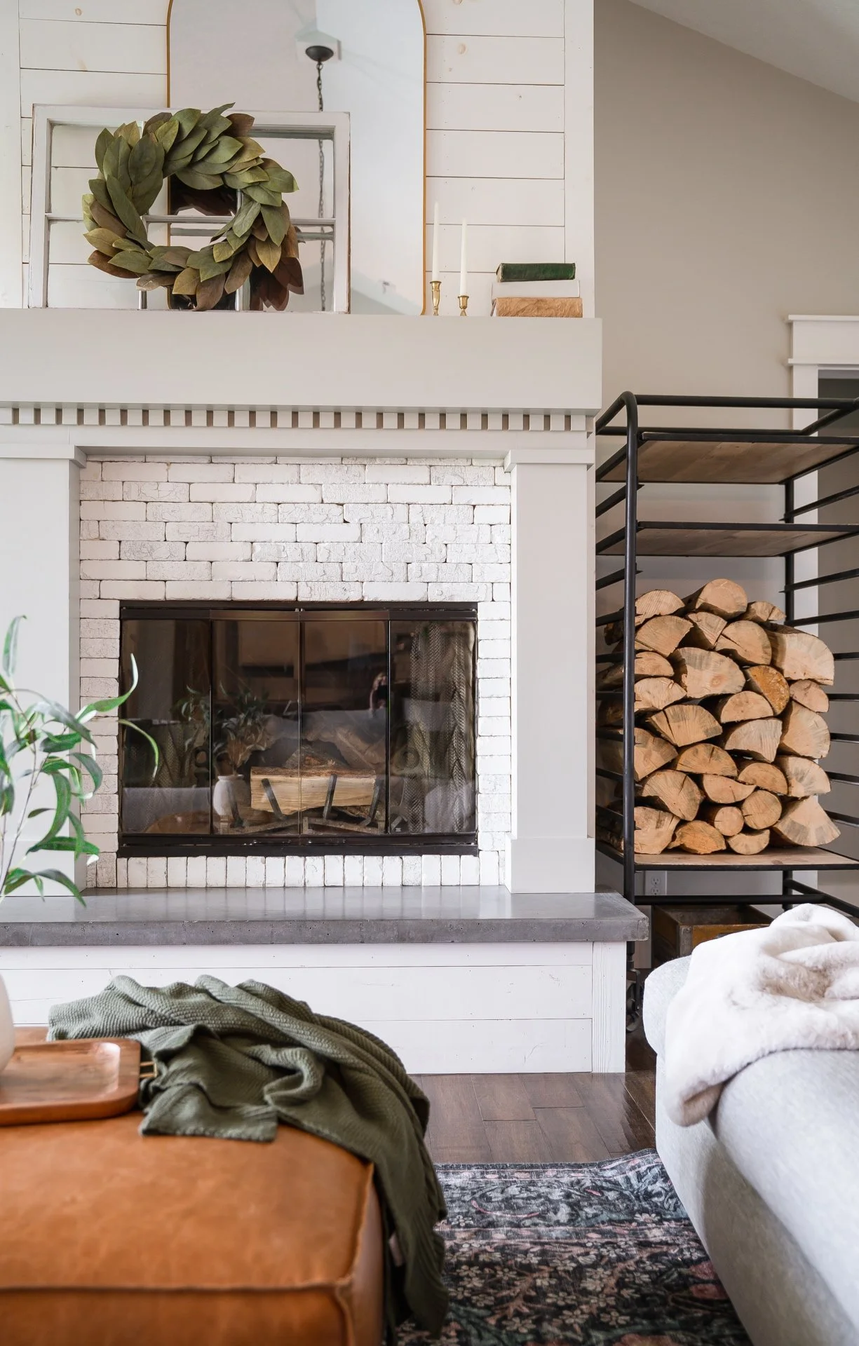 Living room with a painted white brick fireplace, stacked firewood on a side rack, a leather ottoman with a blanket, and a light gray sofa in front.