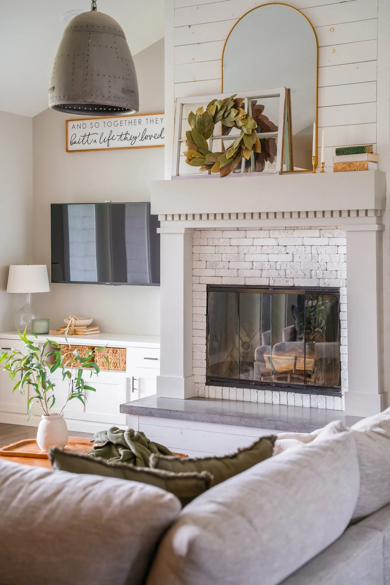 Living room with white brick fireplace, wall-mounted TV, beige couch with pillows, potted plant, and decorative items on the mantle including a wreath and books. A framed quote sign hangs above the TV. A large gray pendant light is overhead.