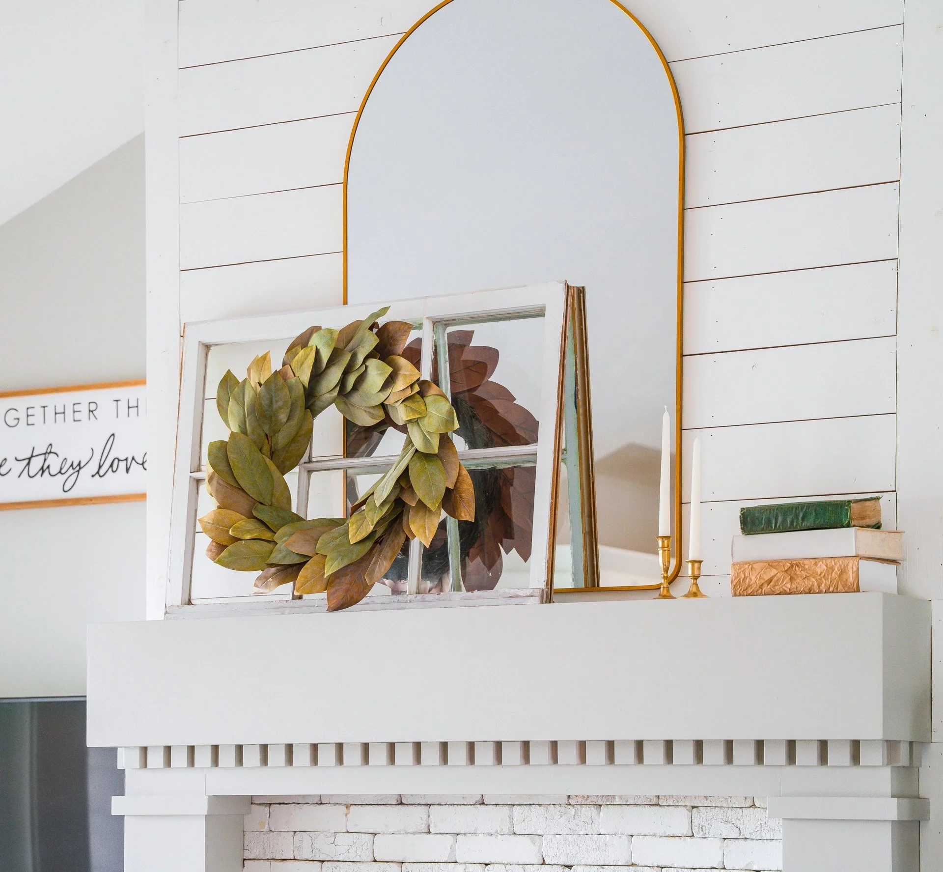Decorative arrangement on a white mantelpiece includes a window frame wreath, a vertical mirror, a pair of tall candles in gold holders, and a stack of books, all set against a white shiplap wall with a framed sign to the left.