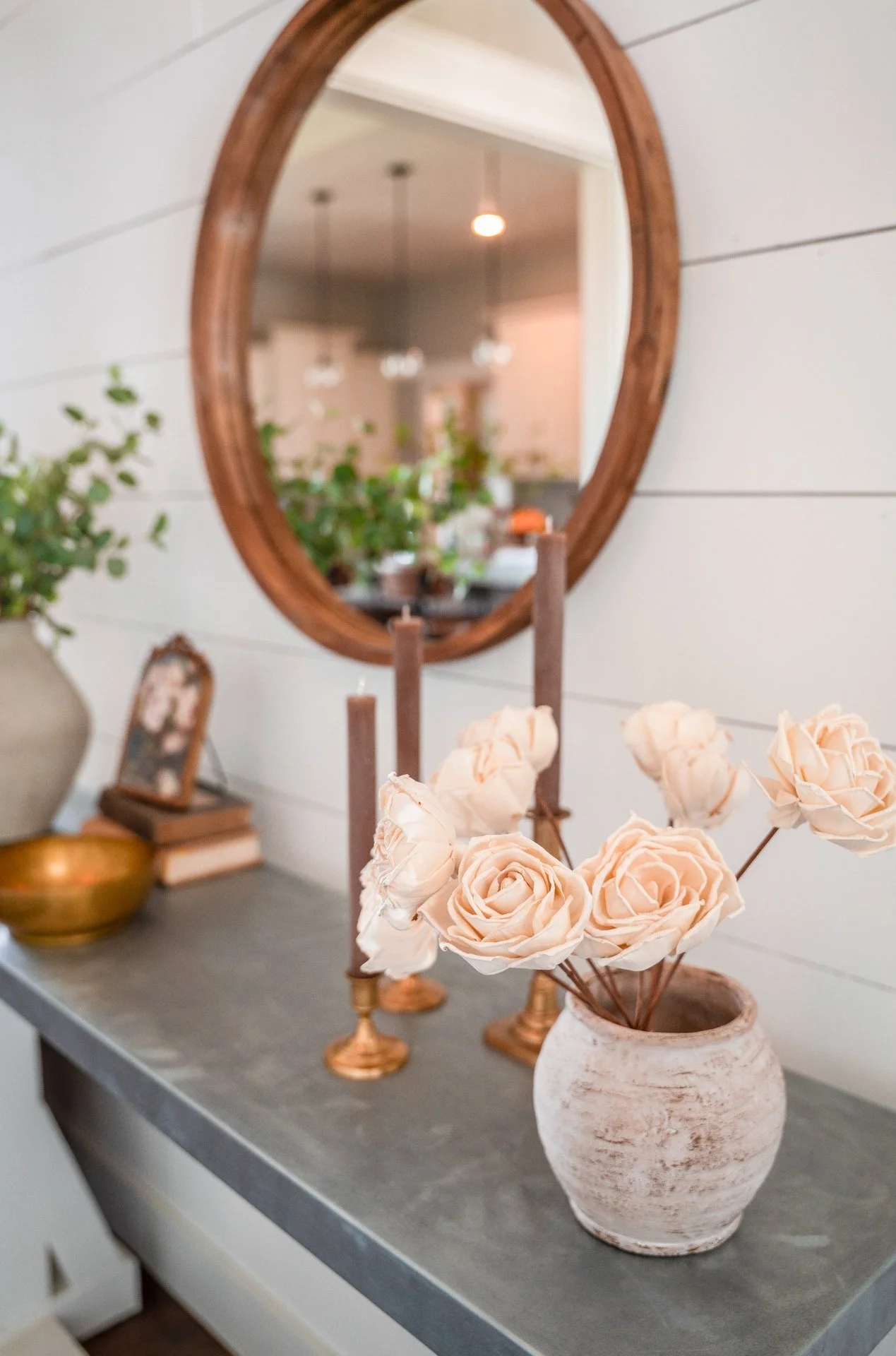 Decorative arrangement on a console table with a beige vase of roses, two tall candles, a bowl, and picture frame of painted flowers, reflected in an oval wooden mirror on a white shiplap wall.