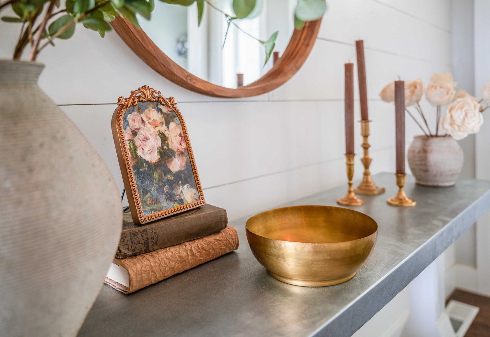 Decorative tabletop display with vases, candlesticks, books, a small framed painting, a wooden bowl, and a flower arrangement in neutral tones and soft lighting.