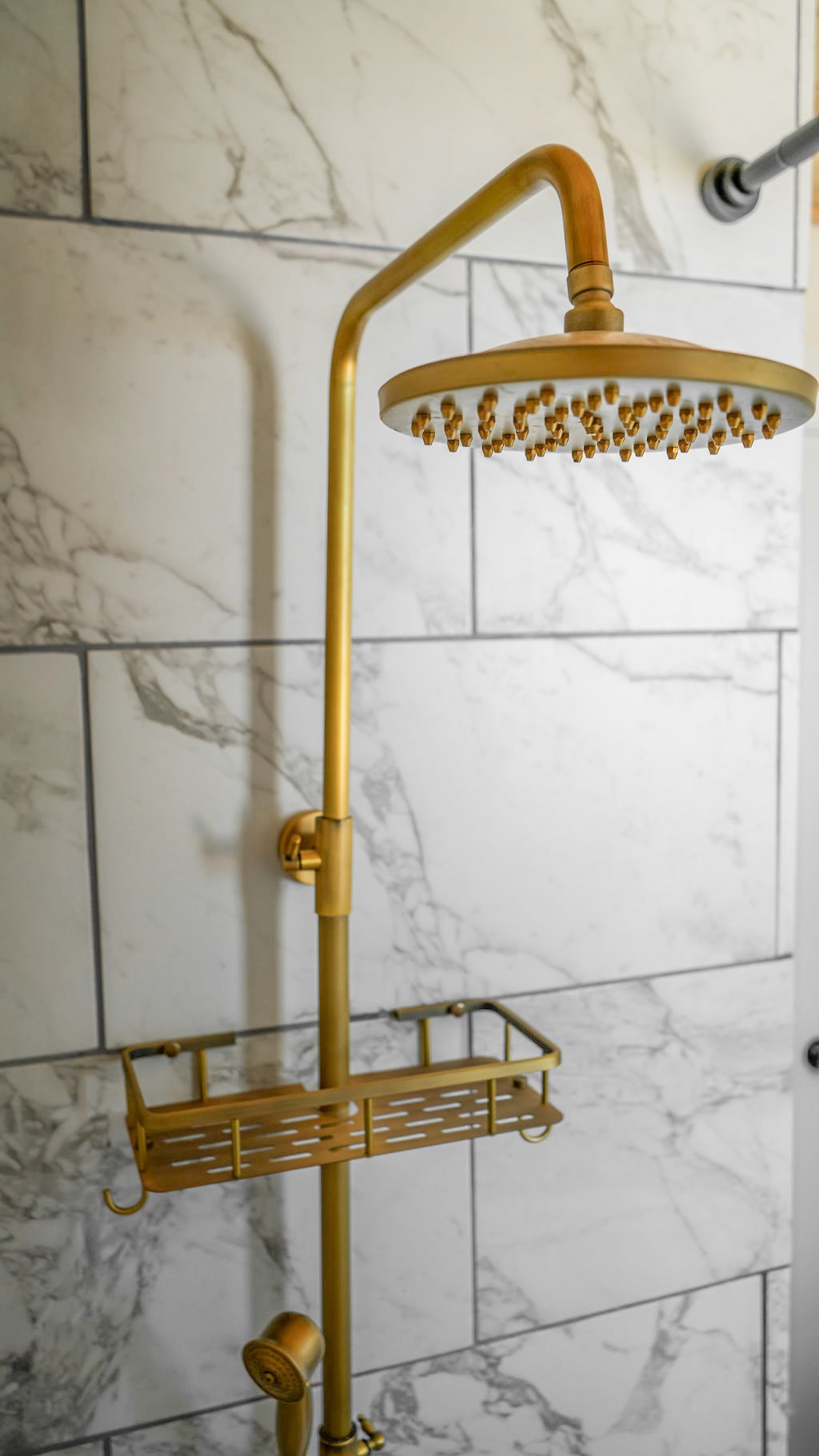 A gold-colored rainfall showerhead with a shower caddy attached, mounted on a wall with large white marble tiles.