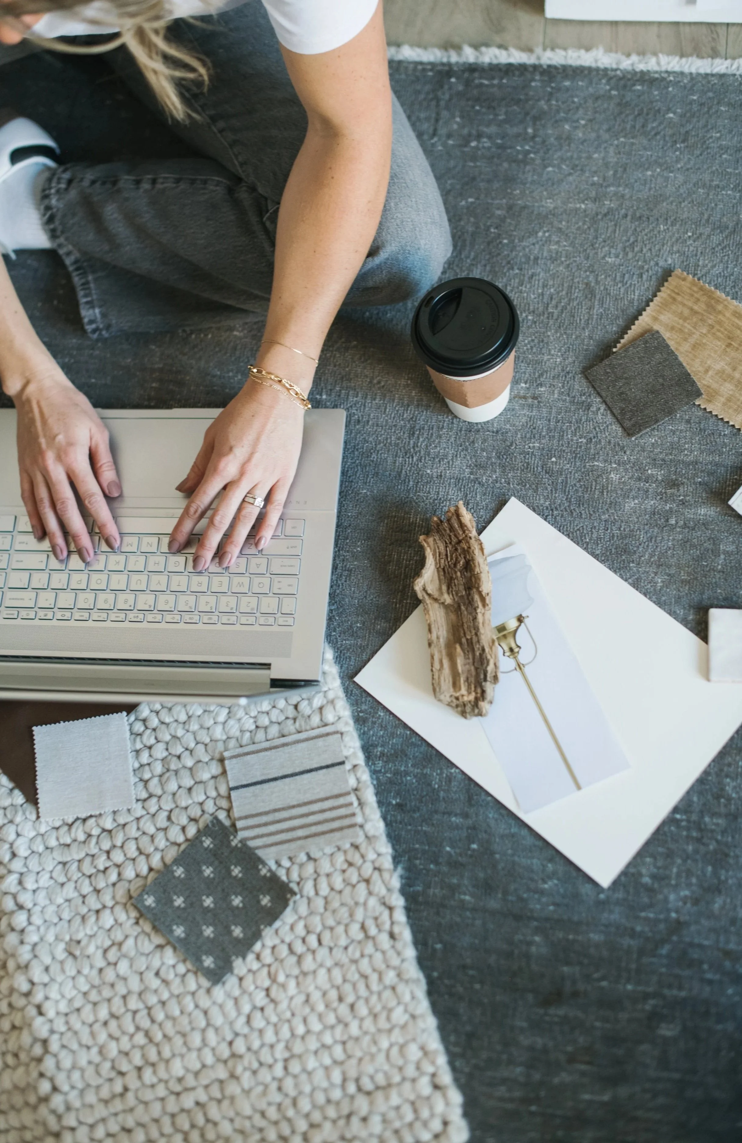 Person working on a laptop surrounded by fabric samples, a coffee cup, and decorative items on a dark textured floor.