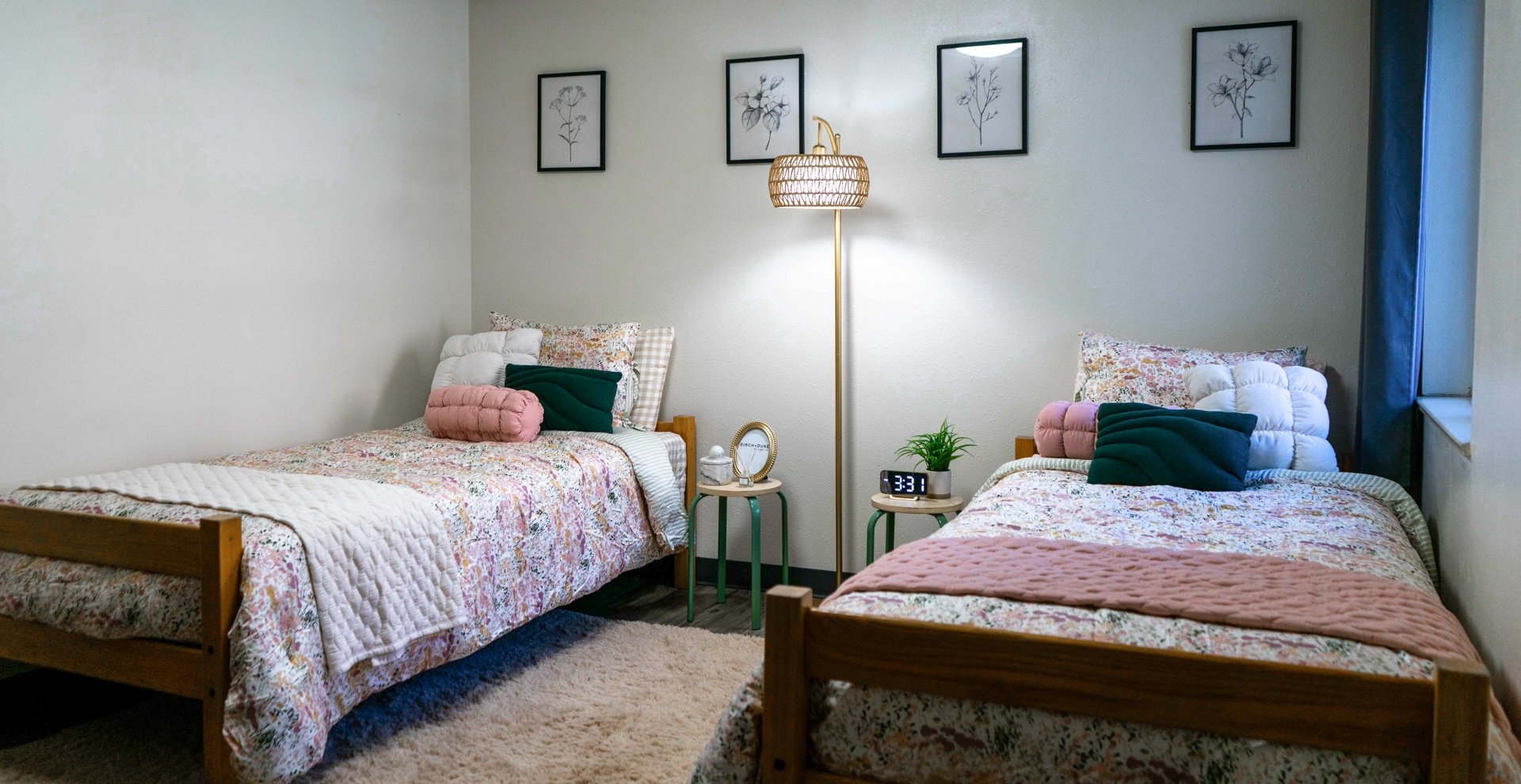 A bedroom with two twin beds with floral bedspreads, pink and green pillows, a wool blanket, and a beige shaggy rug. There is a nightstand between the beds with a plant, a clock, and a small decorative item. A tall floor lamp with a woven shade sits between the beds. Above the beds are four framed botanical prints on a gray wall.