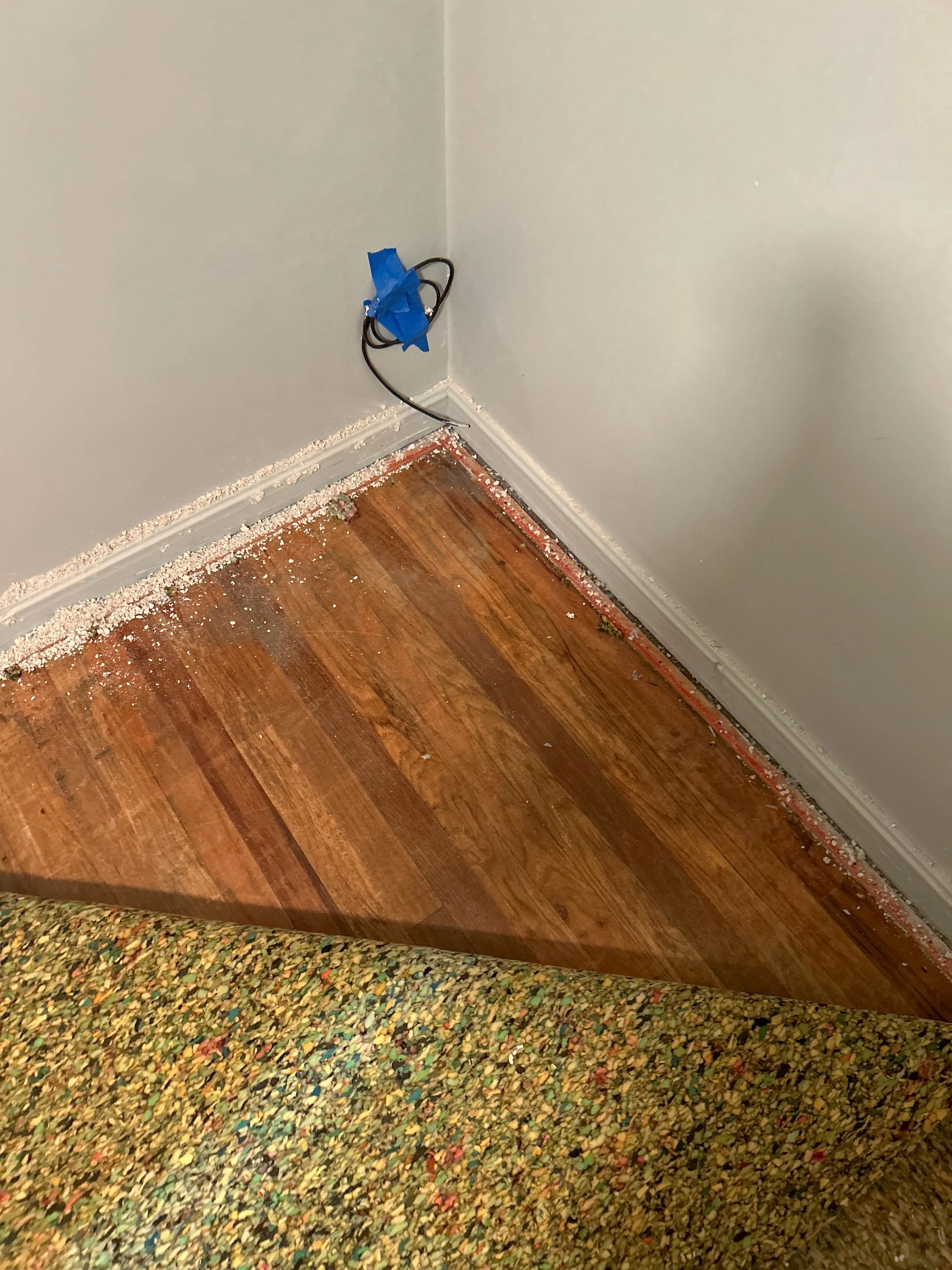 Corner of a room showing a section of hardwood floor, a wall with baseboard trim, debris on the floor, and a black cable taped to the wall with blue painter's tape.