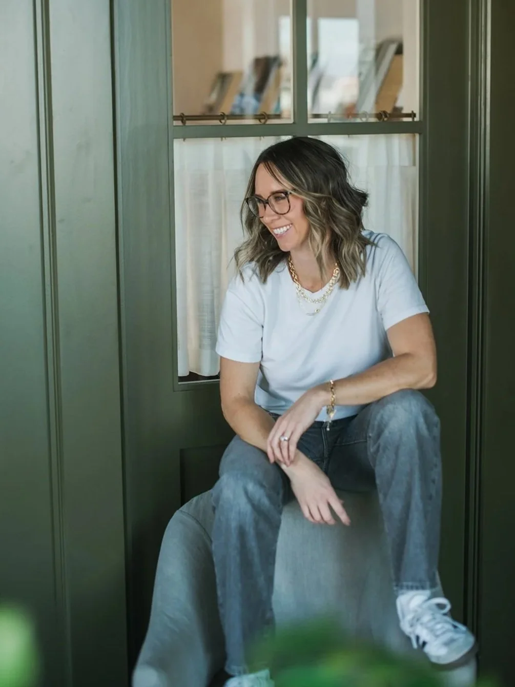 A portrait of interior designer, Trista Cross. She has shoulder-length wavy hair, glasses, wearing a white t-shirt and jeans, sitting on a cushioned seat by a green window, smiling.