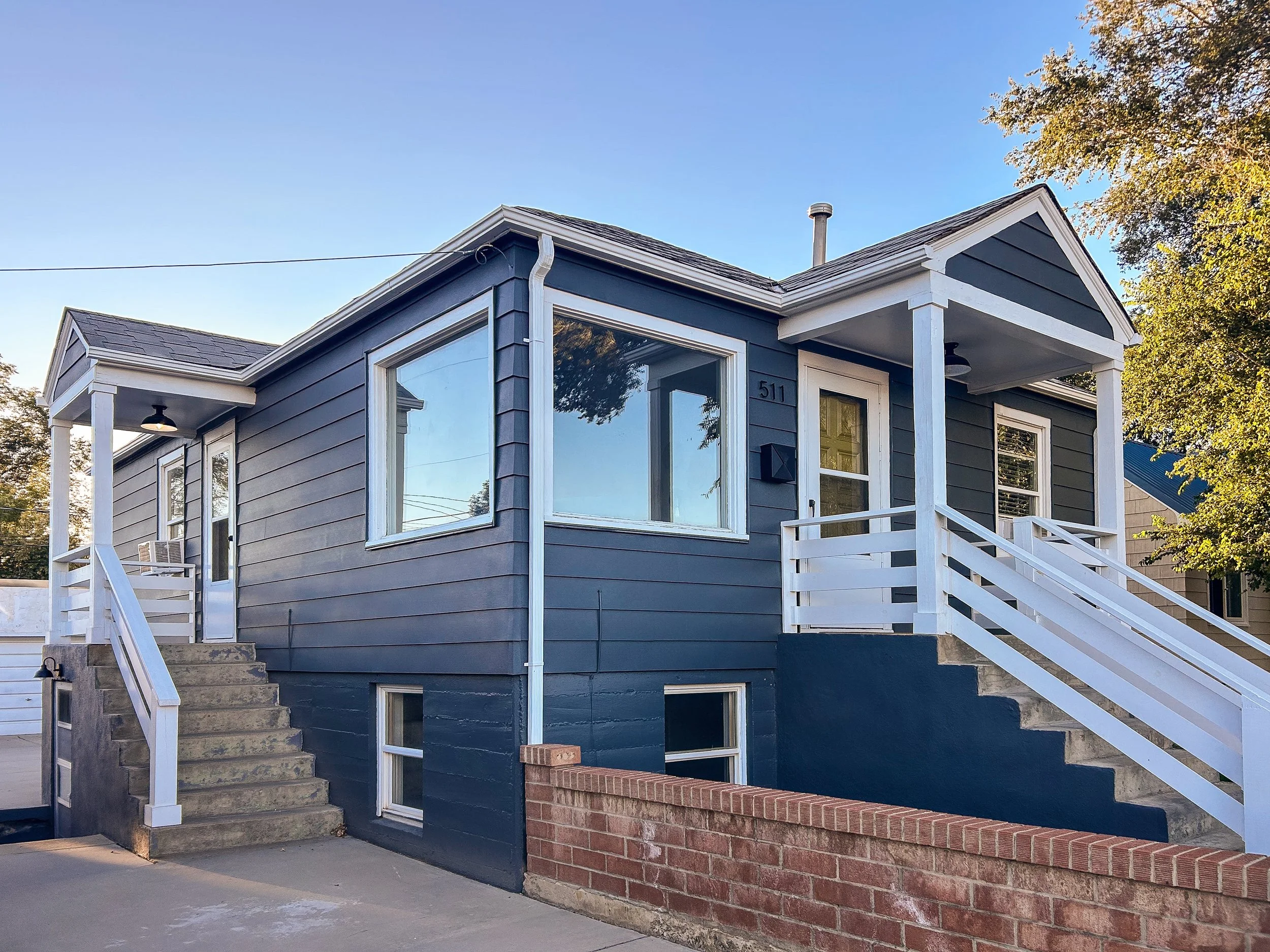 A blue two-story house with white trim and a small porch with stairs leading to the front door. The house has large windows, a brick foundation, and is surrounded by trees.