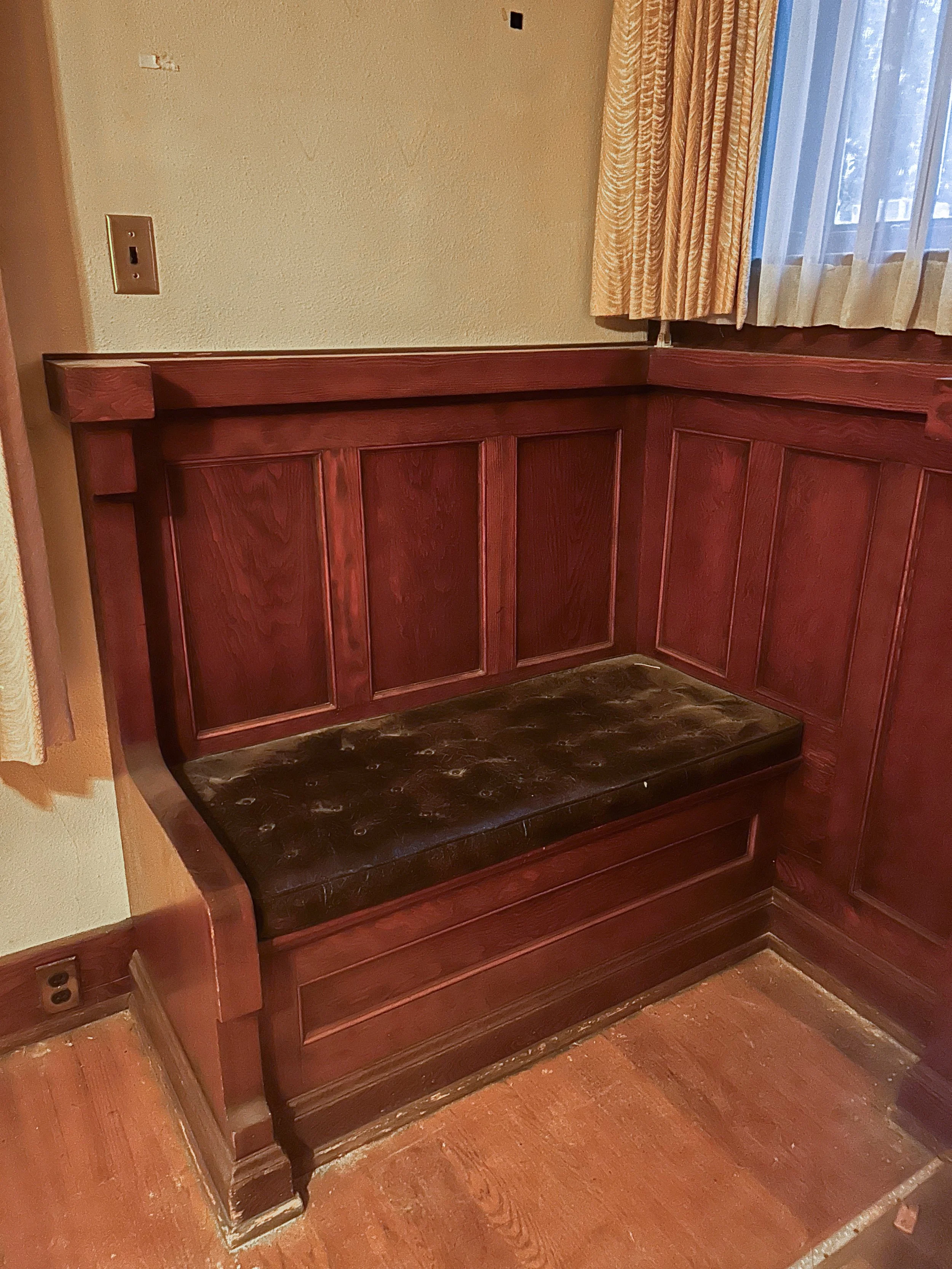 Corner built-in wooden bench with a dark leather cushion in a room with beige walls, wooden floors, and a window with yellow curtains.