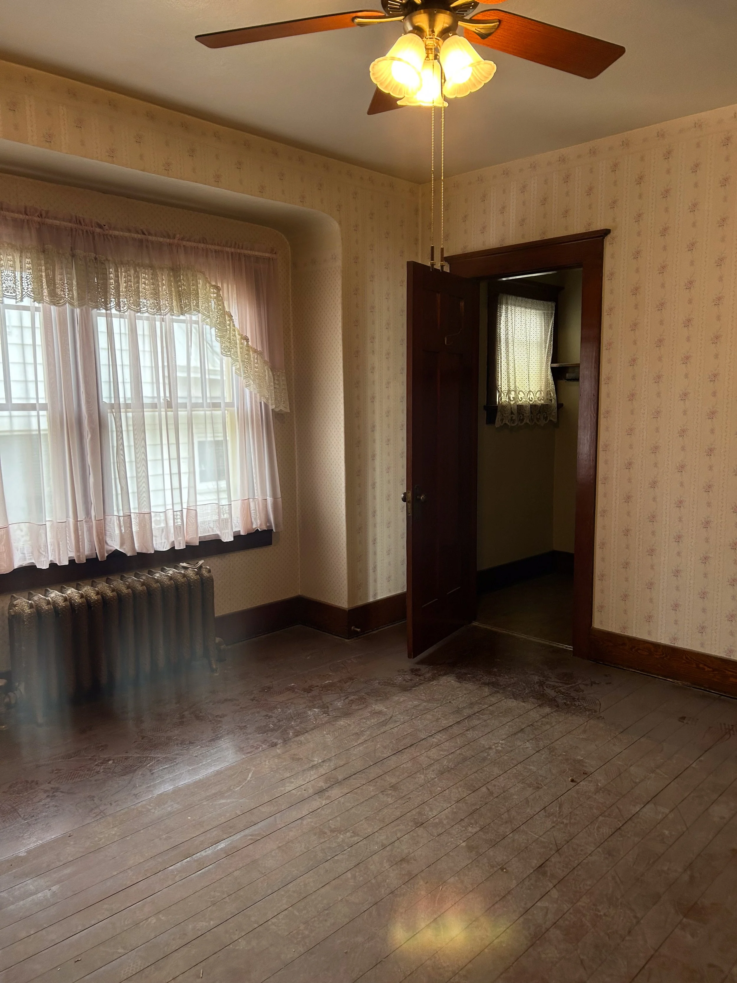 An empty room with wooden flooring, floral wallpaper, a window with pink curtains, a ceiling fan with lights, and a partially open wooden door leading to a small closet space.