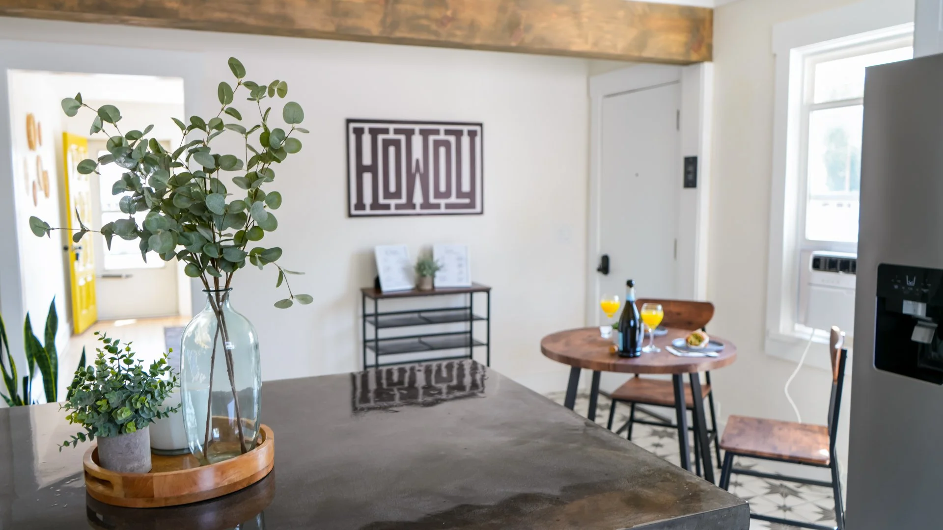 Kitchen with pendant lighting, potted plants, a durable concrete countertop, a round wooden table set for breakfast with a large window and "Howdy" framed art on the wall.