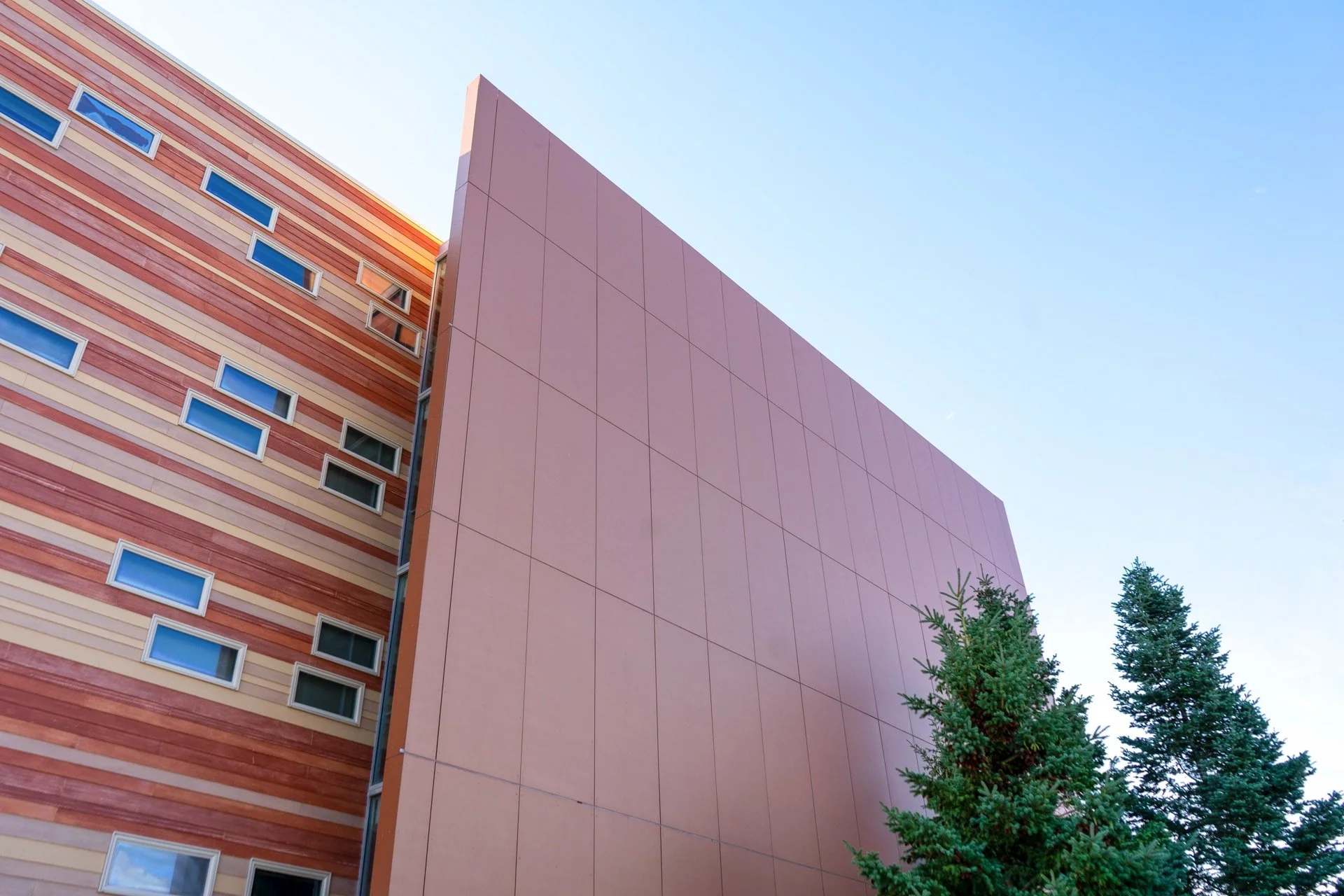 Low-angle view of a modern building with colorful horizontal bands and small rectangular windows, next to a plain pink wall and trees, against a clear blue sky.