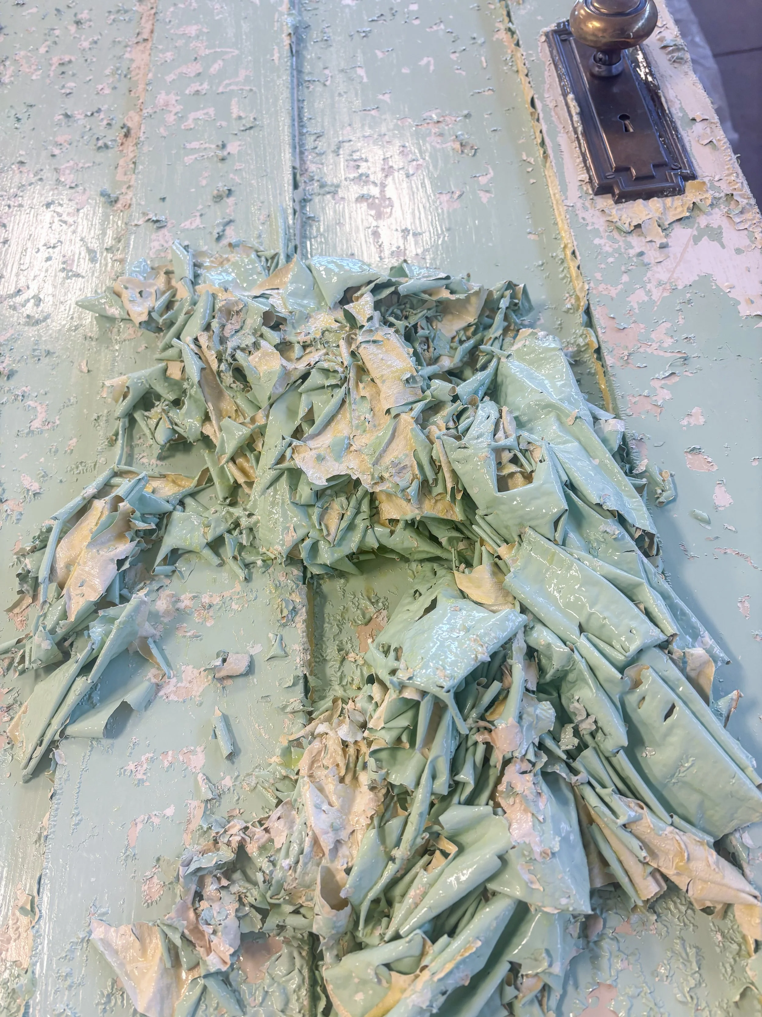 An antique green painted door with peeling paint and a pile of paint that has been scraped off of the door sitting in front of it.