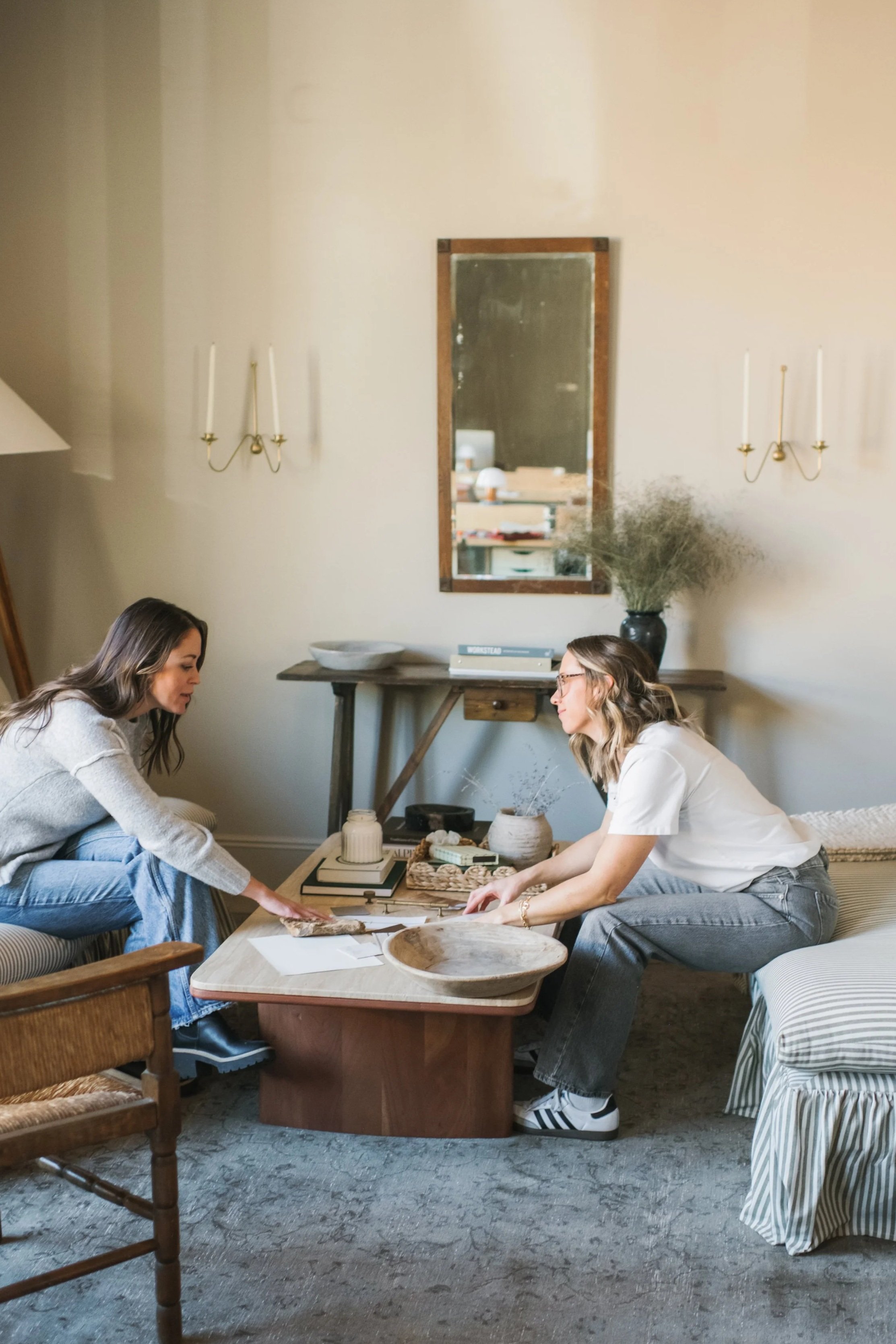 Two women are sitting in a living room, engaged in a conversation at a coffee table with decorative items, a tray, and papers. One woman is on the left, sitting on the edge of a sofa, and the other is on the right, leaning forward. There is a wooden console table with books and a large vase with dried plants behind them. The room has soft lighting and a mirror on the wall.