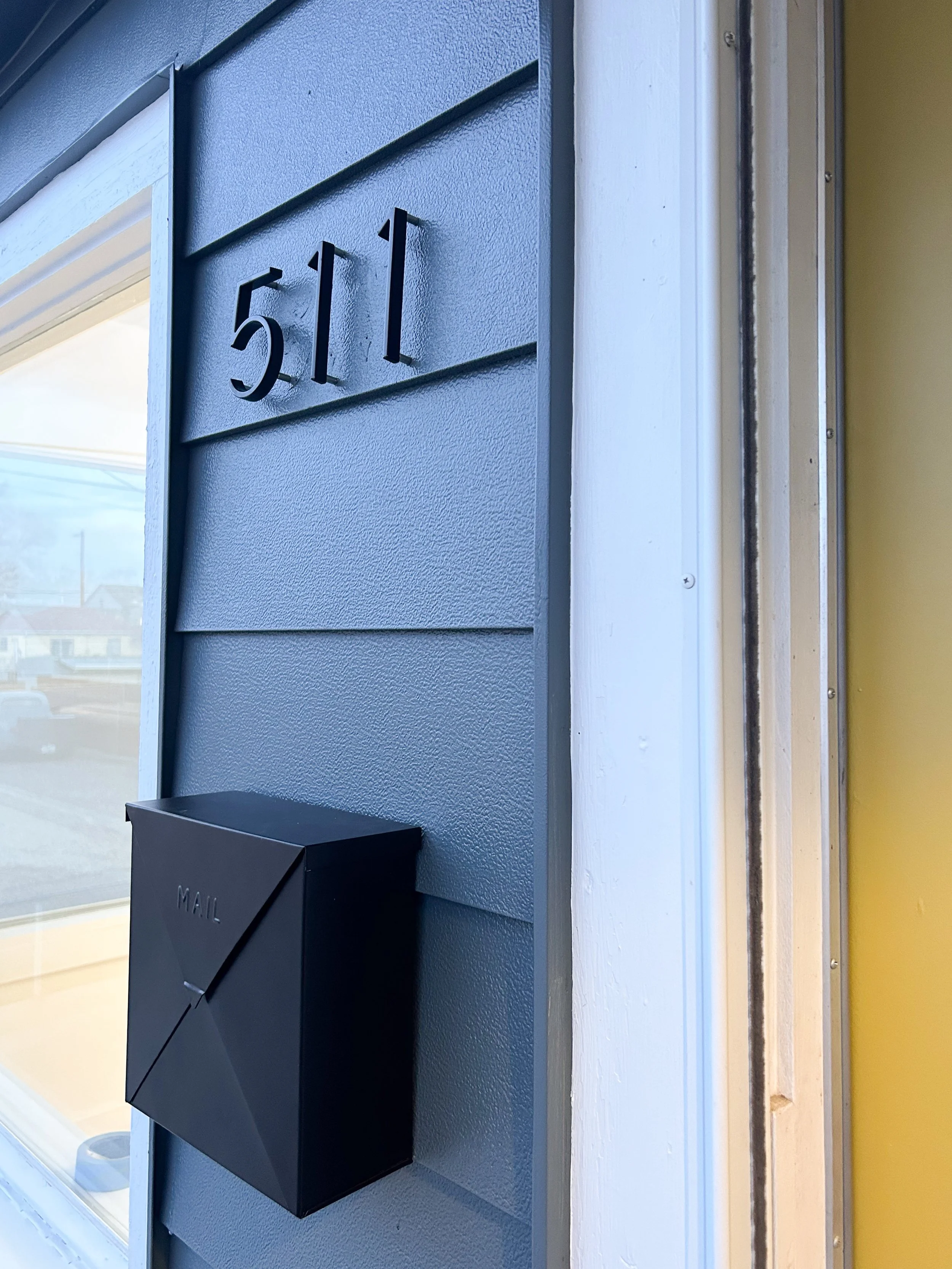 Close-up of a gray house exterior with the address 51 underneath black, three-dimensional numbers, and a black mailbox labeled 'MAIL' mounted on the wall beside a window. The house has horizontal siding and a white trim, with part of a yellow wall visible on the right.