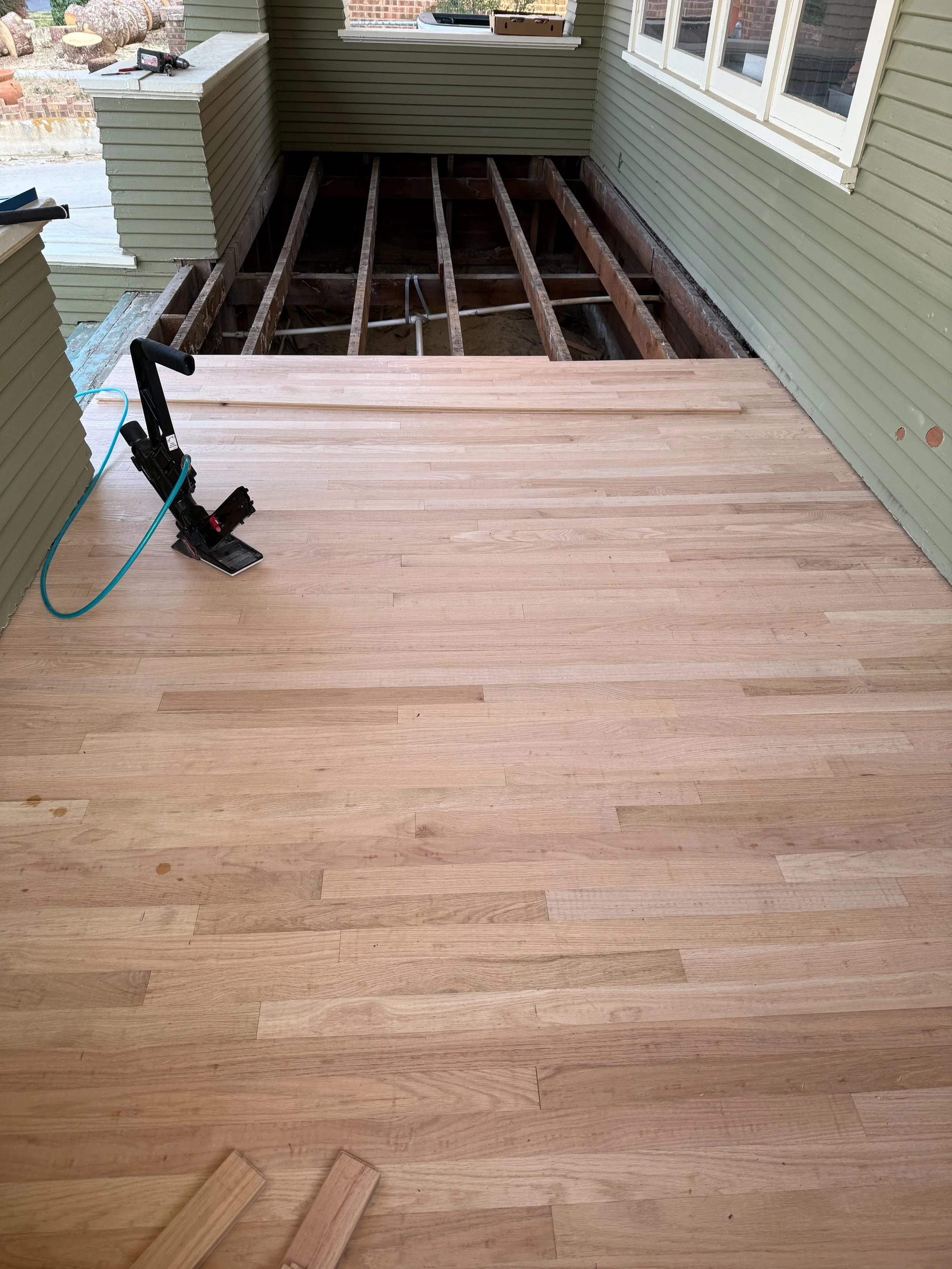 A partially completed hardwood floor installation on a front porch, with an electric sander and some wood planks on the floor, and the unfinished area of the porch exposing exposed floor joists.