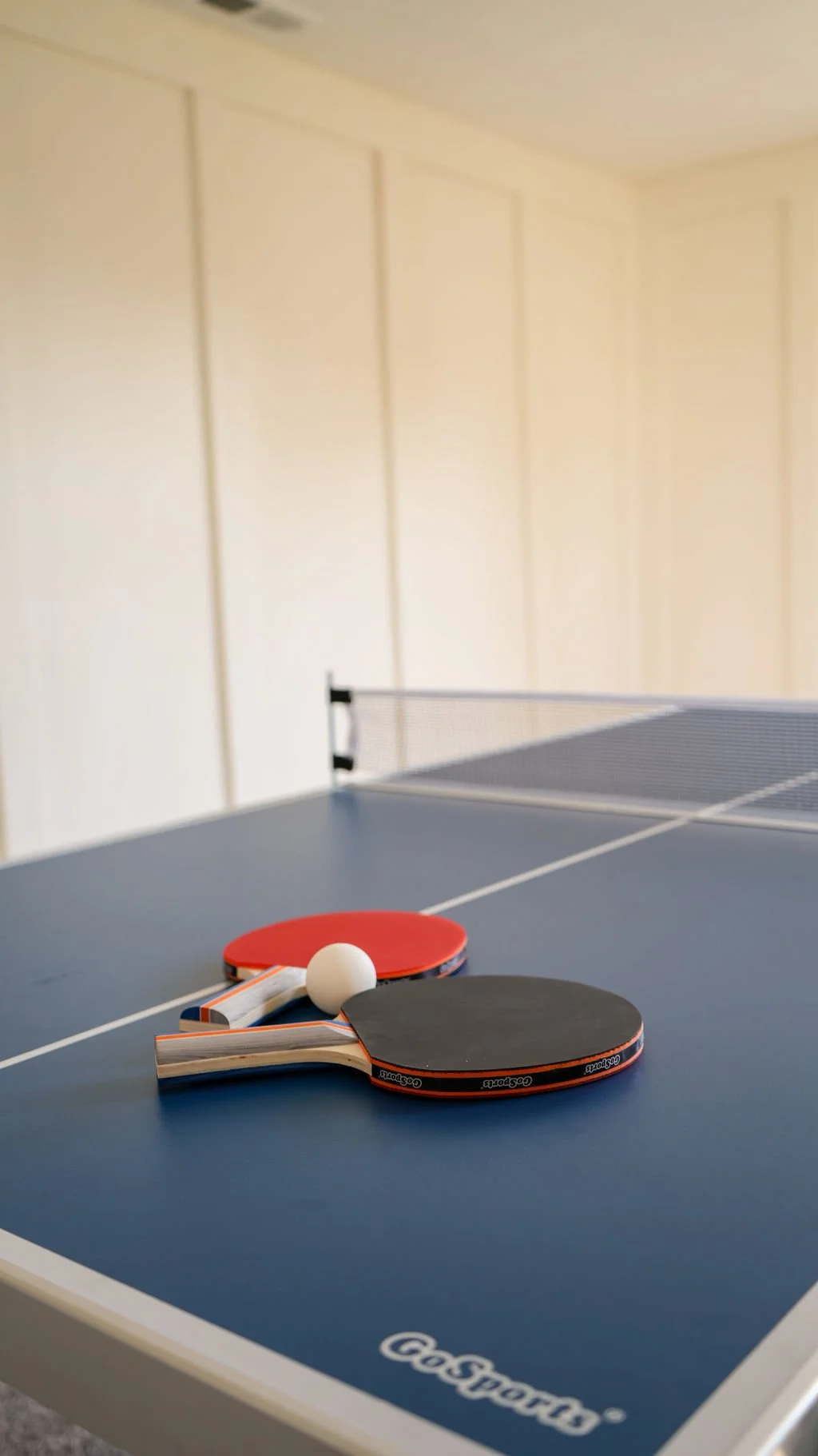 A blue table tennis table with two paddles, a ball, and a net. The paddles are red and black, with the black paddle labeled 'Caspo'. The table is indoors, with cream-colored panel walls in the background.