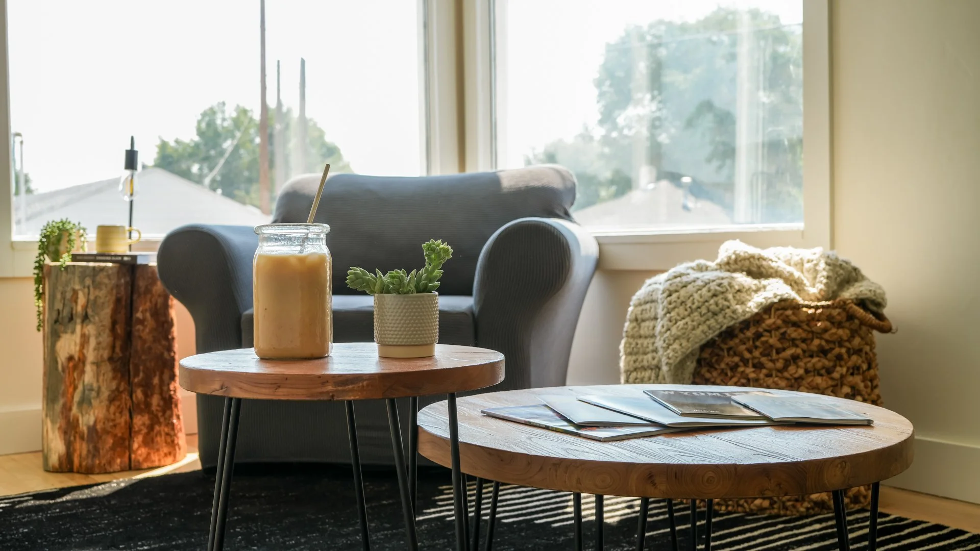 Living room with a black armchair, a small round wooden coffee table with magazines, and a smaller wooden table with a mason jar drink and a small potted succulent, near a large window letting in natural light.