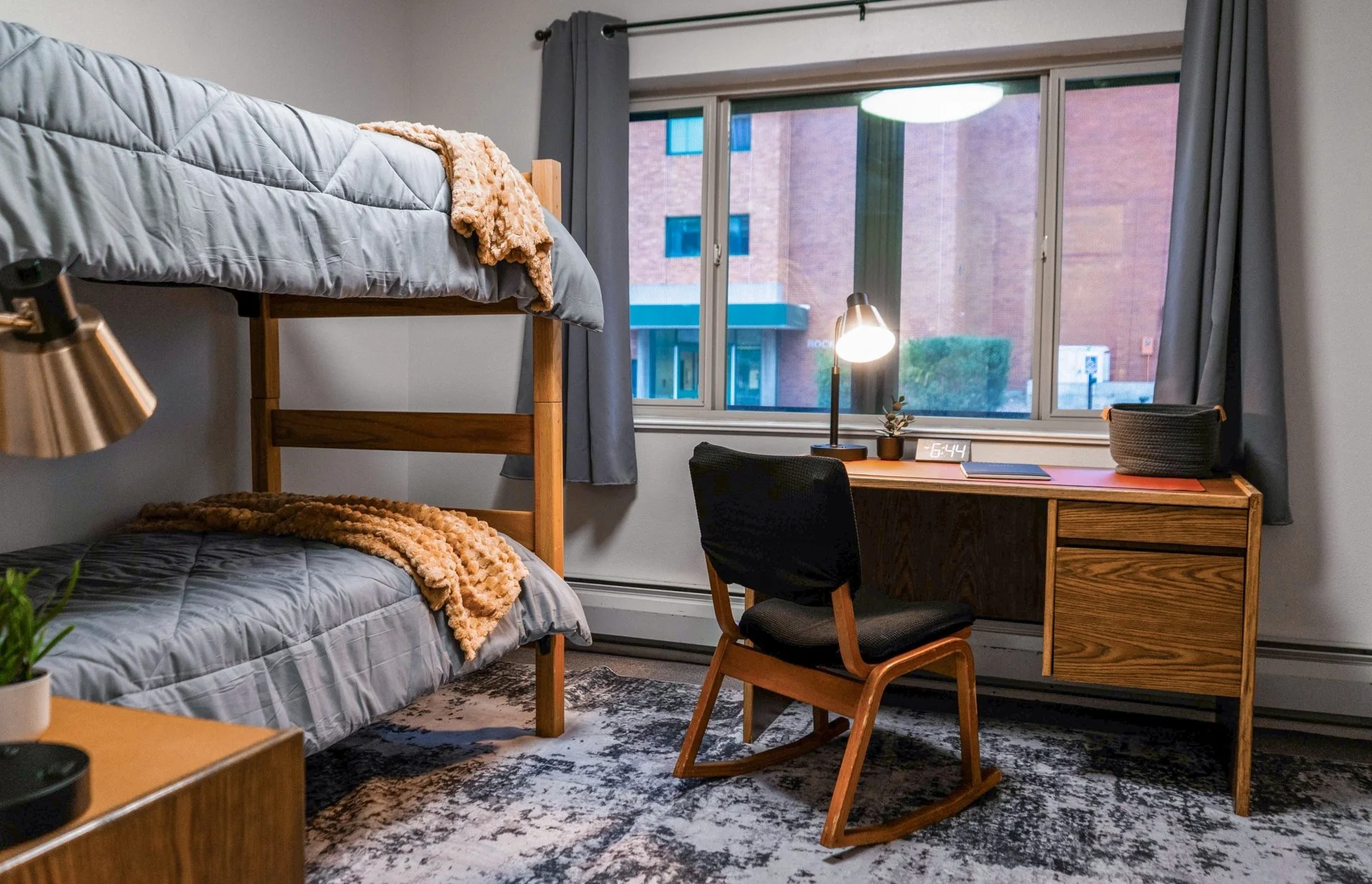 A bedroom with a wooden bunk bed, a desk by the window, a black chair, a lamp, a digital clock, and some plants and storage baskets. The room has dark curtains and a patterned rug.