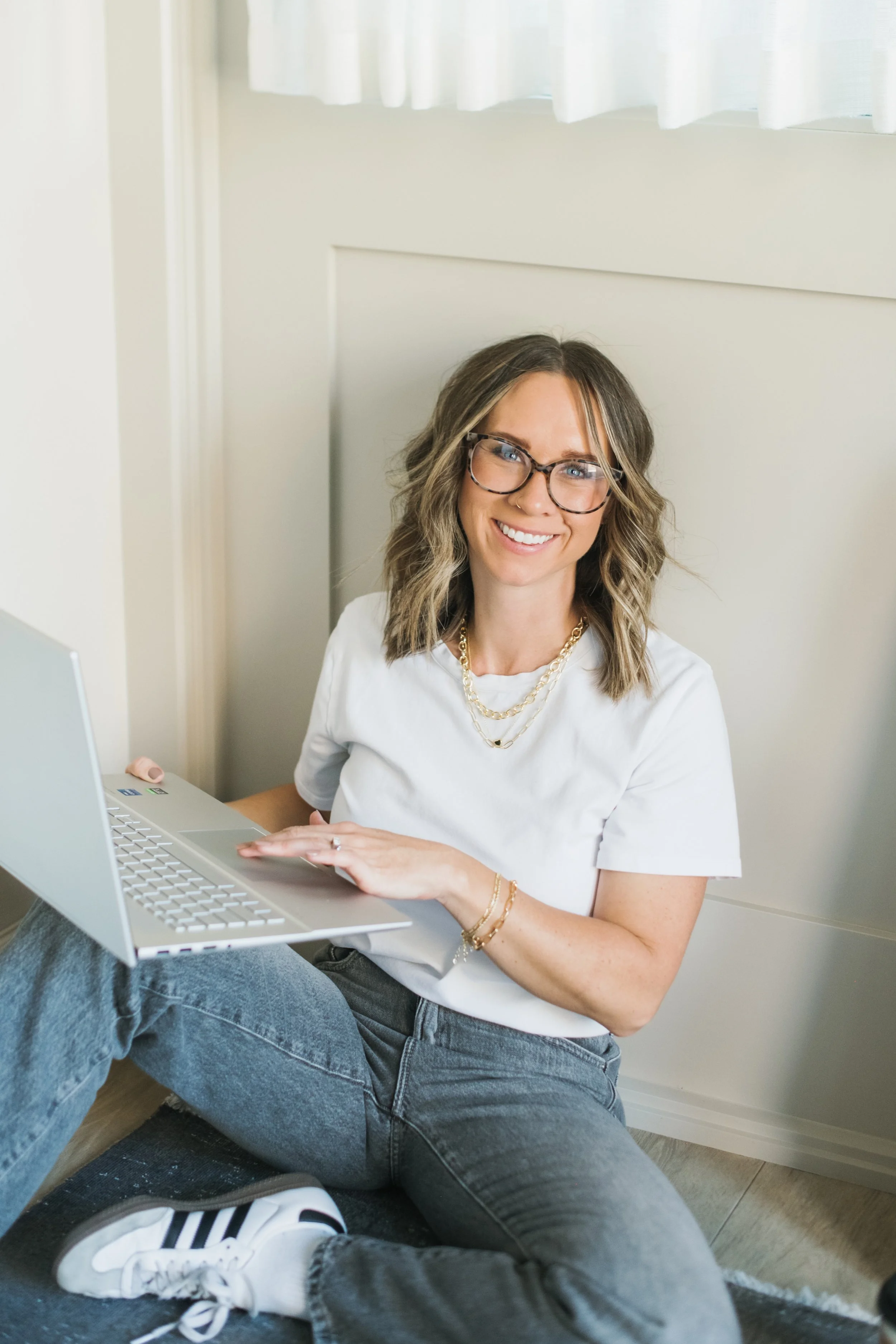 A portrait of interior designer, Trista Cross, wearing glasses and wavy hair sitting on the floor with a laptop in her lap, smiling at the camera.