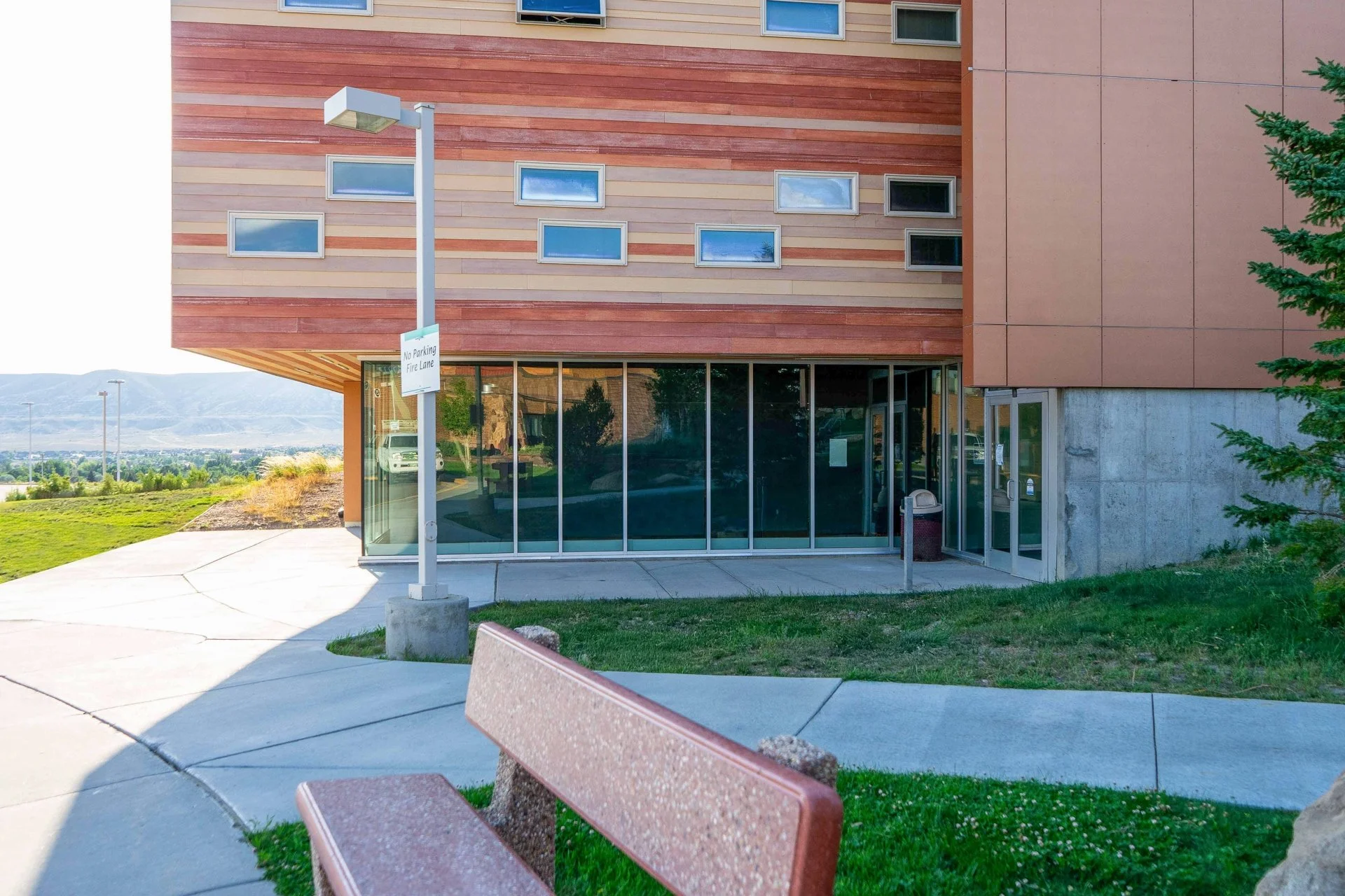 Exterior view of a modern building with large glass windows, a wood-paneled upper section, and a concrete lower section, surrounded by a sidewalk, green grass, and a bench, with a mountain landscape in the background.