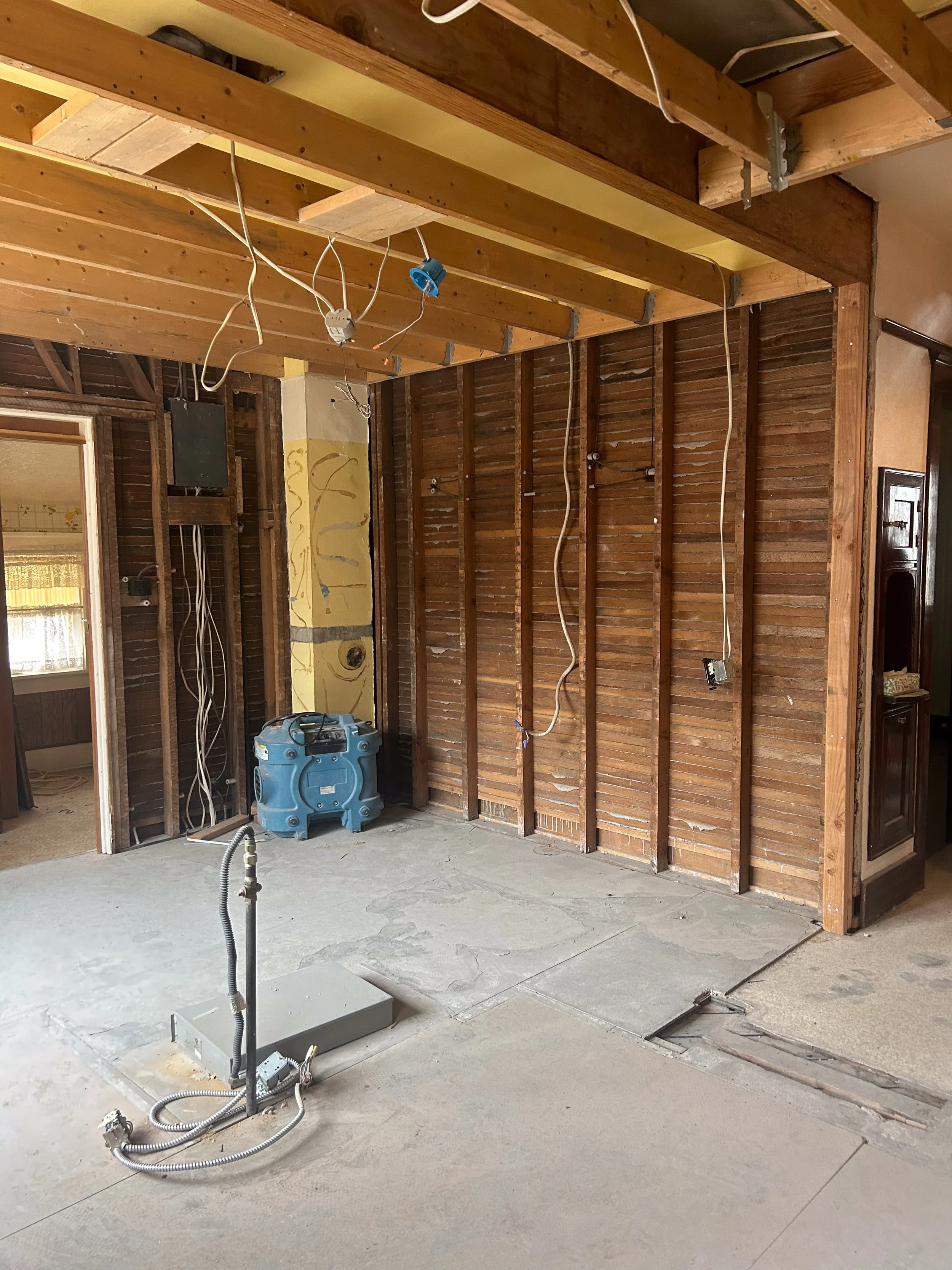 Room under renovation with exposed wooden ceiling beams, exposed wall studs, and electrical wiring hanging from the ceiling. The floor is partially torn up, and a blue air purifier is near the wall.