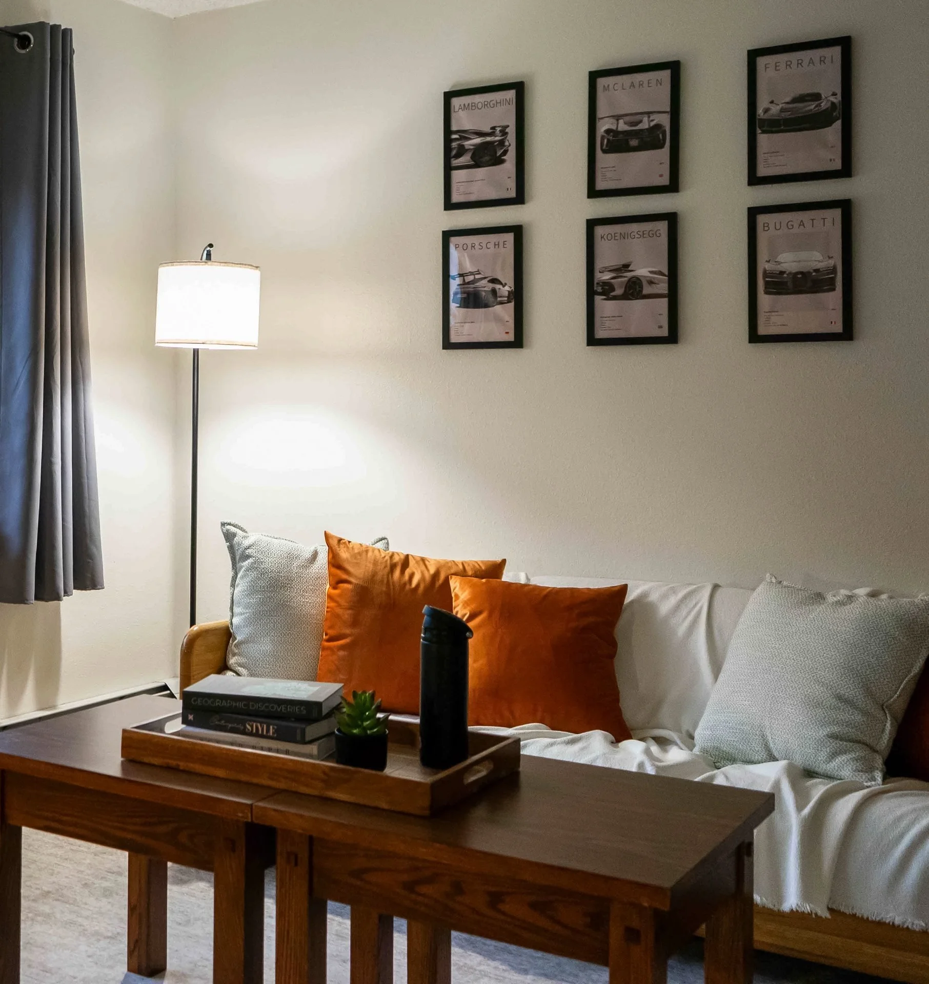 Living room with a white sofa decorated with orange and cream-colored pillows, a wooden coffee table with books, a small plant, and a black thermos, a tall standing lamp, grey curtains, and framed pictures of luxury car brands on the wall.