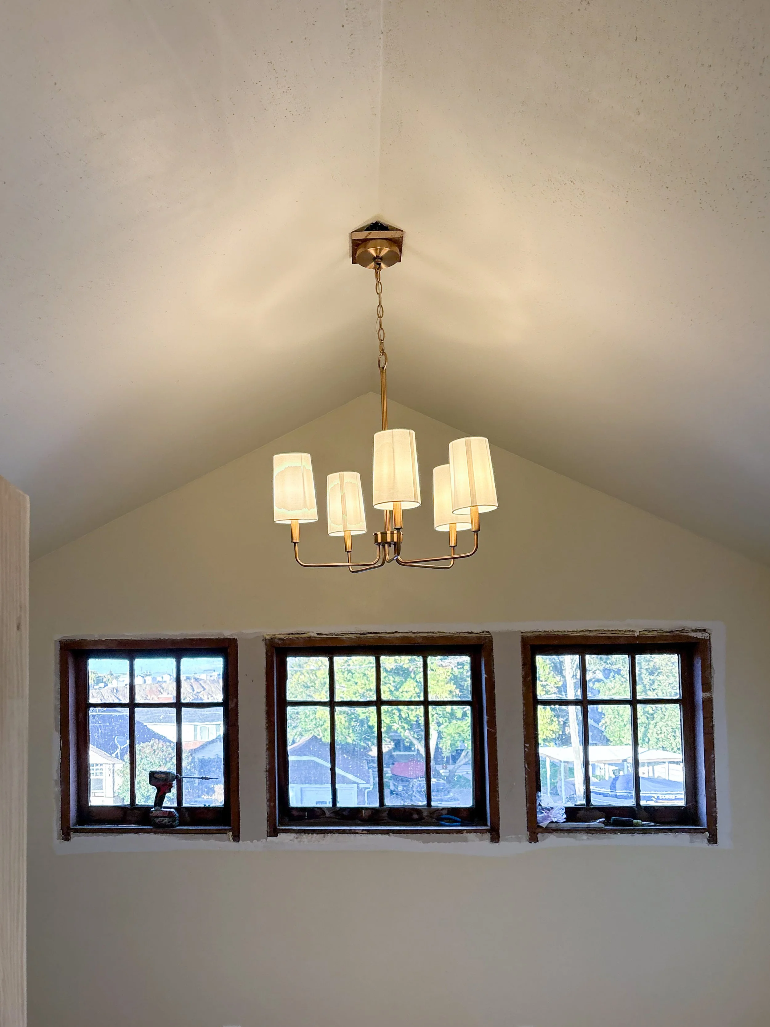 Interior view of a room with a gold chandelier hanging from a chain, three small windows with black grid frames, and visible construction or renovation work around the window frames.
