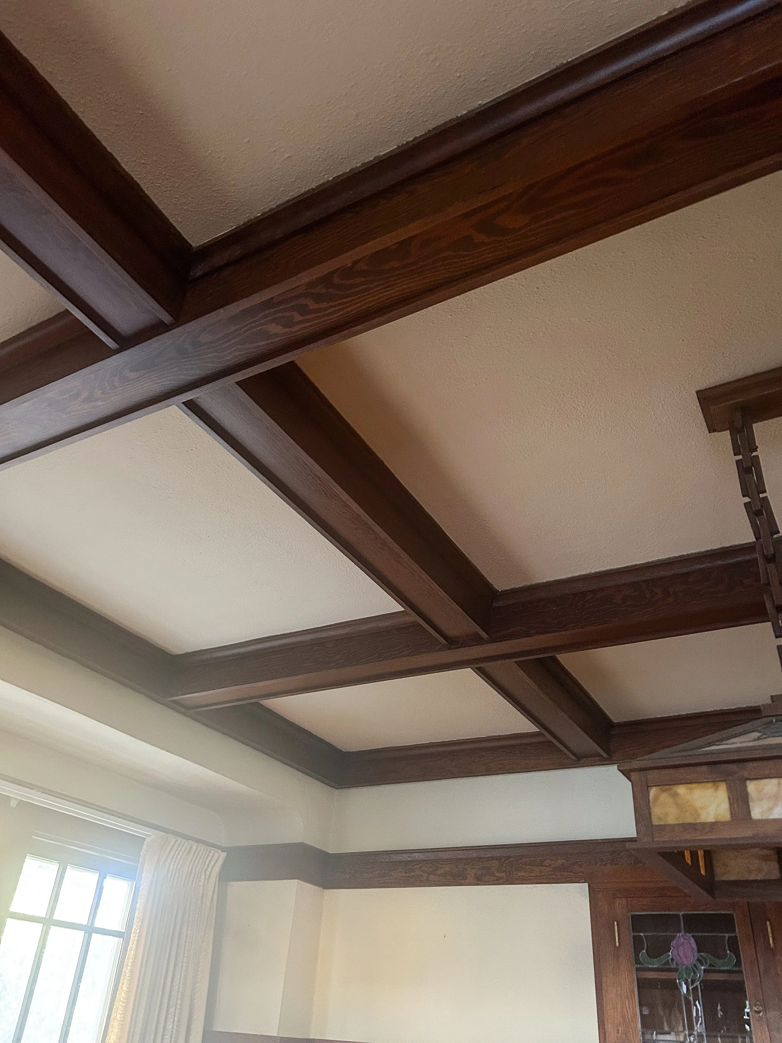 Ceiling with wooden beams and panels in a room with a window and curtains.