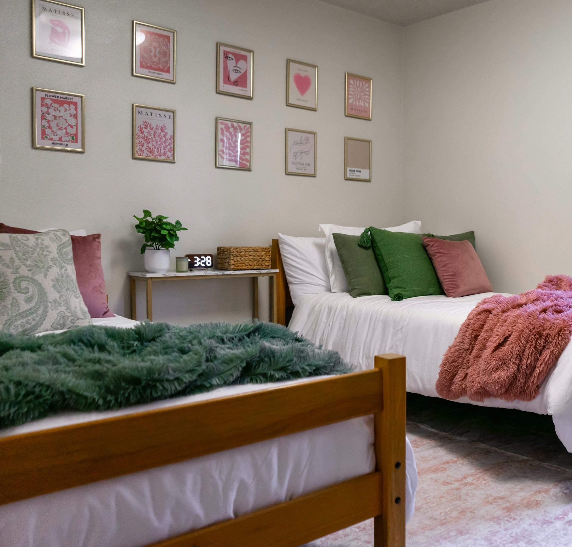 Two beds in a bedroom with wall art, pillows, and blankets, and a nightstand with a plant, wicker basket, digital clock, and cup, with neutral-colored walls and a patterned rug.