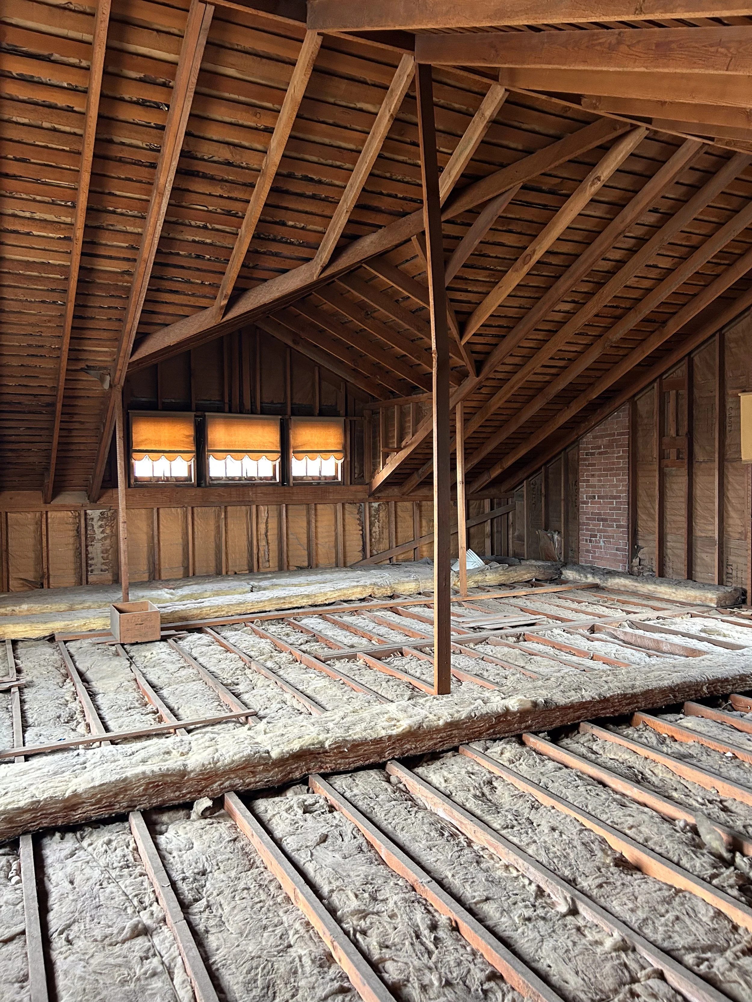 An attic with exposed wooden beams and insulation on the floor, with a small window covered by a yellow blind.
