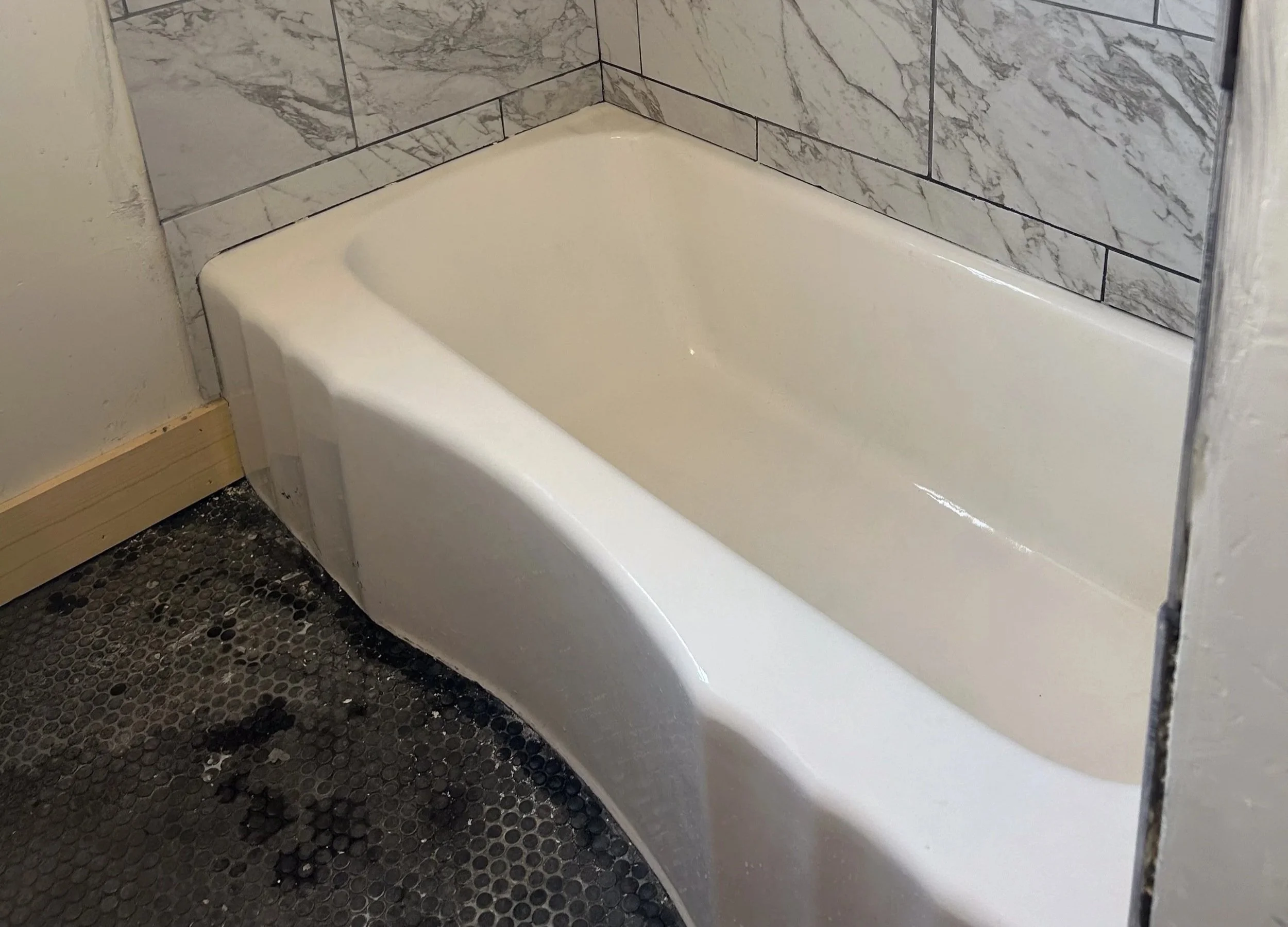 Scalloped edge vintage bathtub with gray and white marble tiled walls and black penny tile floors.