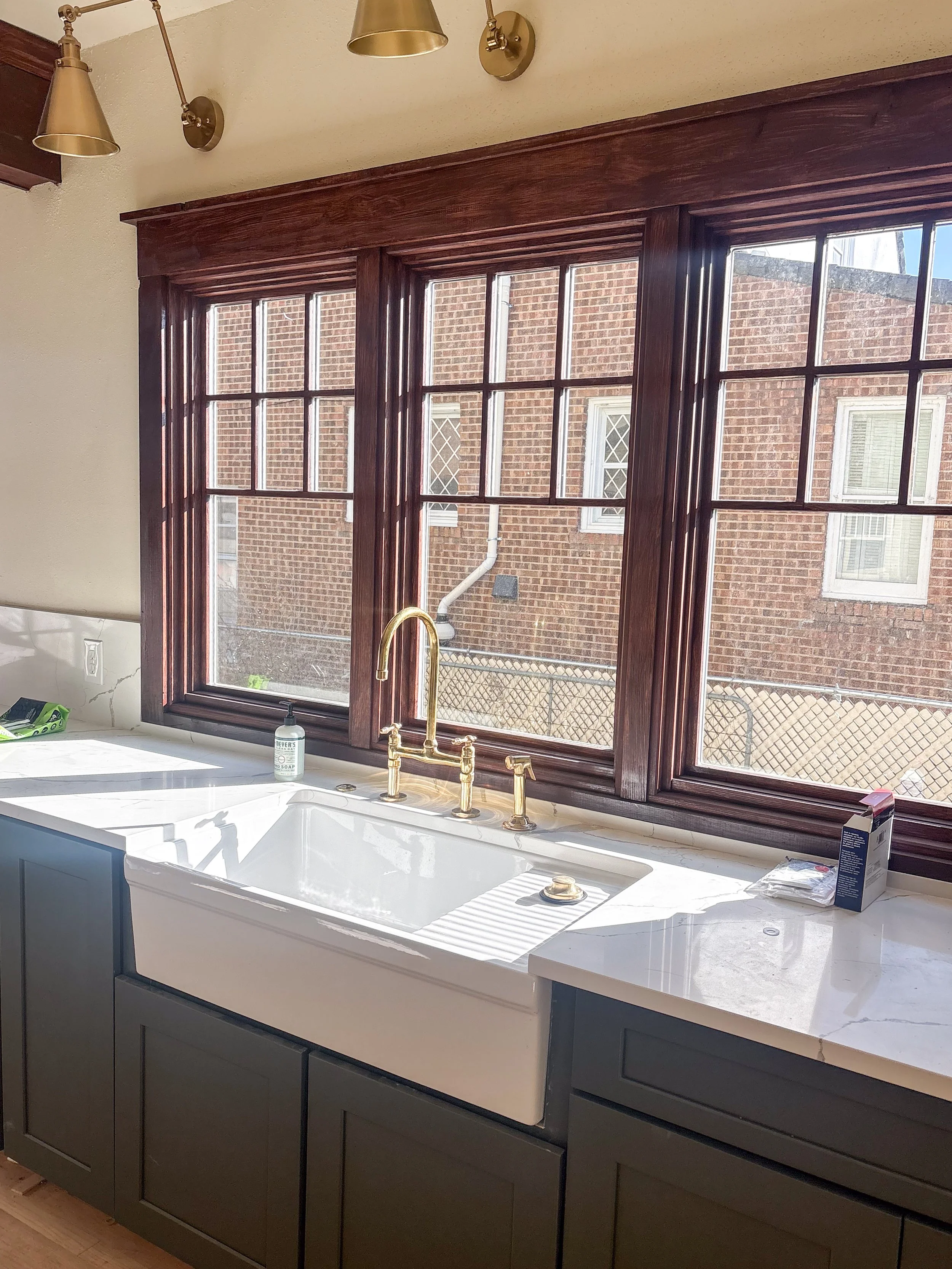Kitchen sink with a white farmhouse style basin, mounted on dark green cabinetry, and brass fixtures. Large window with wooden trim allows sunlight to fill the space, with an outside brick wall visible through the window.
