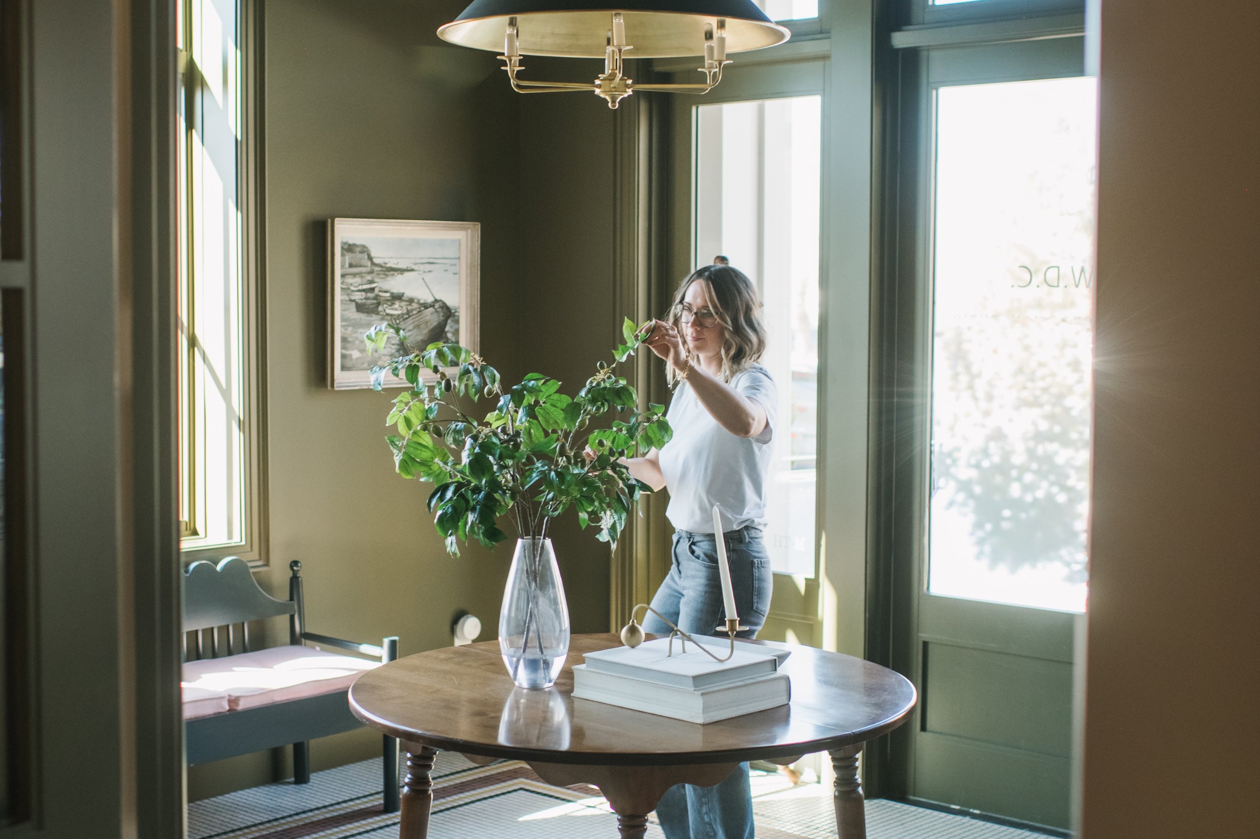 A portrait of interior designer, Trista Cross arranging a plant in a glass vase on a wooden table in a sunlit room with green walls.