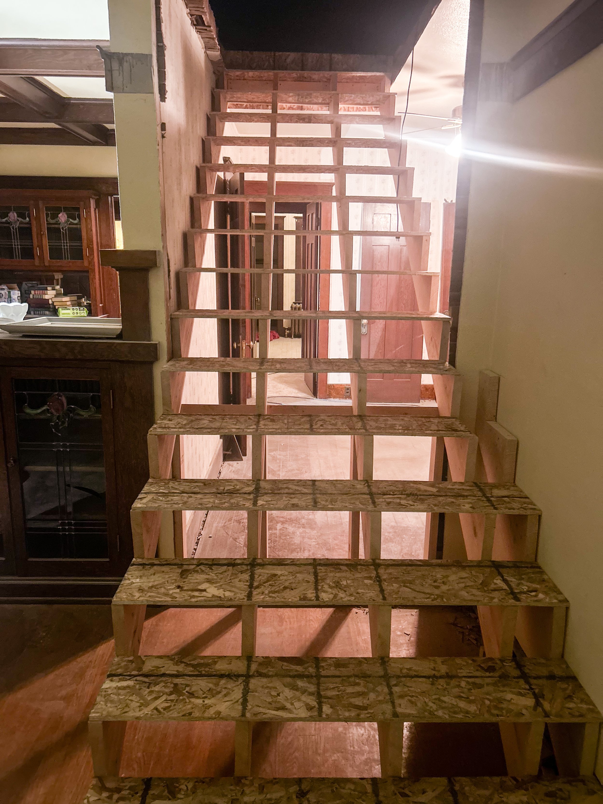 The image shows a staircase under construction with wooden framing. The stairs are unfinished, with no treads or handrails, leading up to a second floor. The surrounding area is part of a home interior with a cabinet and dining area visible on the left.