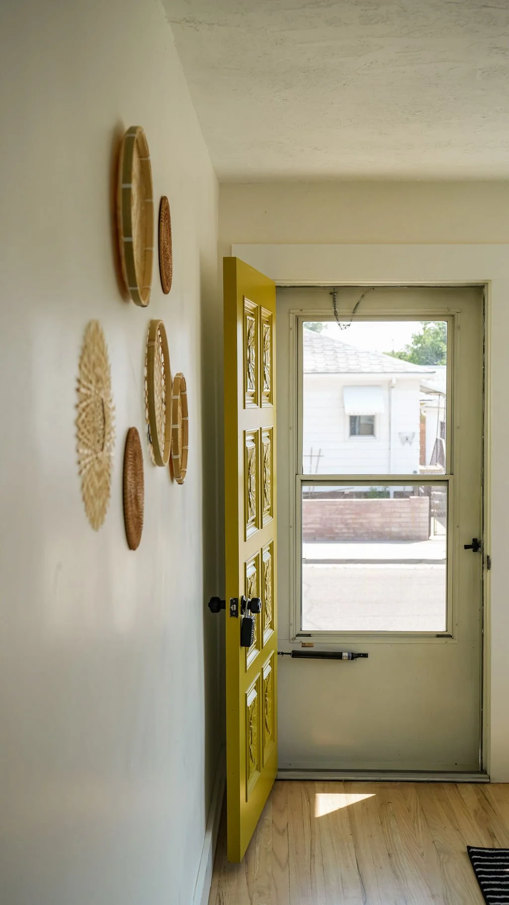 Yellow front door with a window, a doorbell, and a lock, open to reveal part of a small porch and neighboring house, sunlight on hardwood floor. Large wicker trivets hang on the wall to the left.