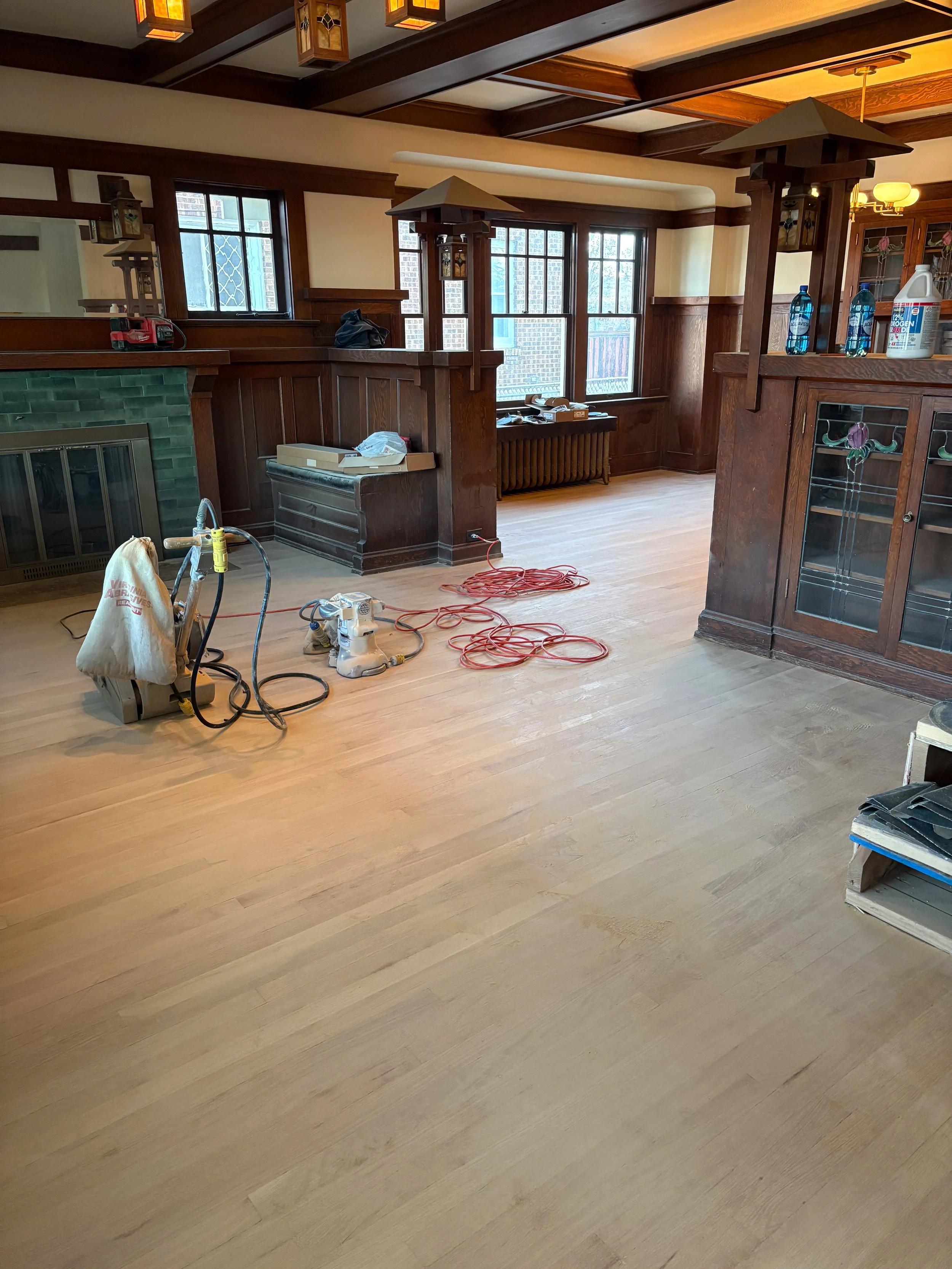 A room under renovation with light wooden flooring, wooden paneling, and large windows. Construction tools and equipment are on the floor, including a vacuum, extension cords, and a small sander. The room has a fireplace with a green tile surround and dark wood trim, and built-in wooden shelves and cabinets.